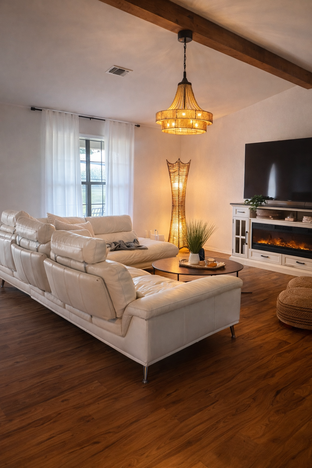 Living room with a beige sectional sofa, a round coffee table with decor, a tall woven floor lamp, a chandelier, a large window with sheer curtains, a white entertainment console with a flat-screen TV, and a fireplace, wooden ceiling beams, and hardw