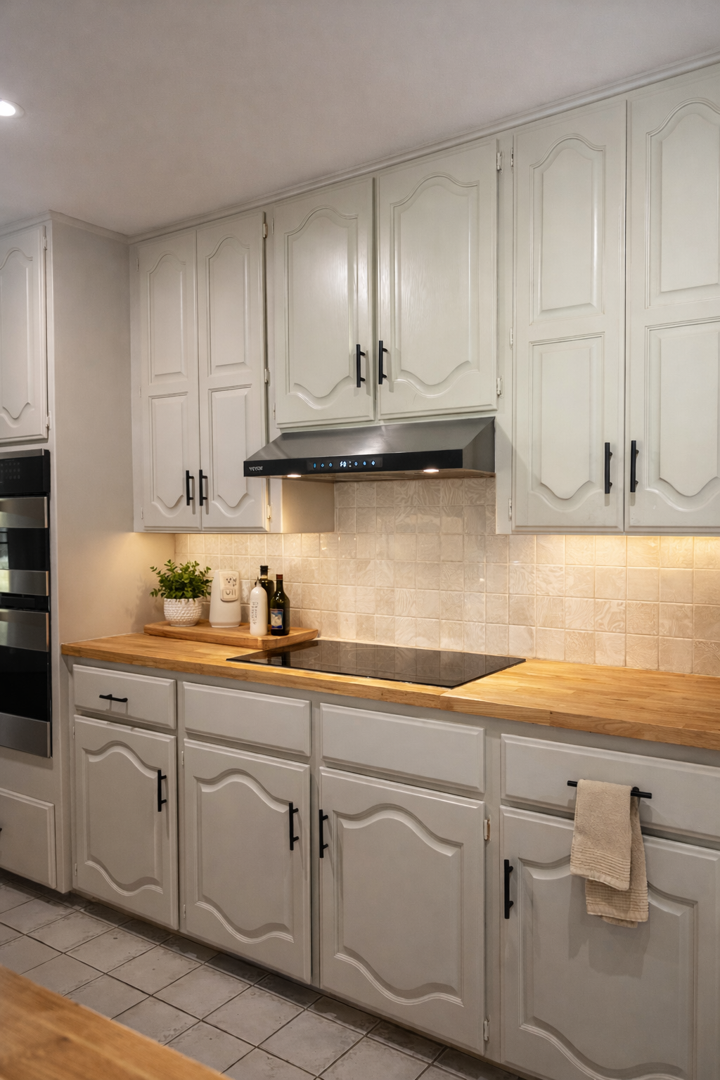 White kitchen cabinets with black handles, wooden countertop, stovetop, range hood, beige tiled backsplash, potted plant, soap dispenser, olive oil bottles, beige towels.