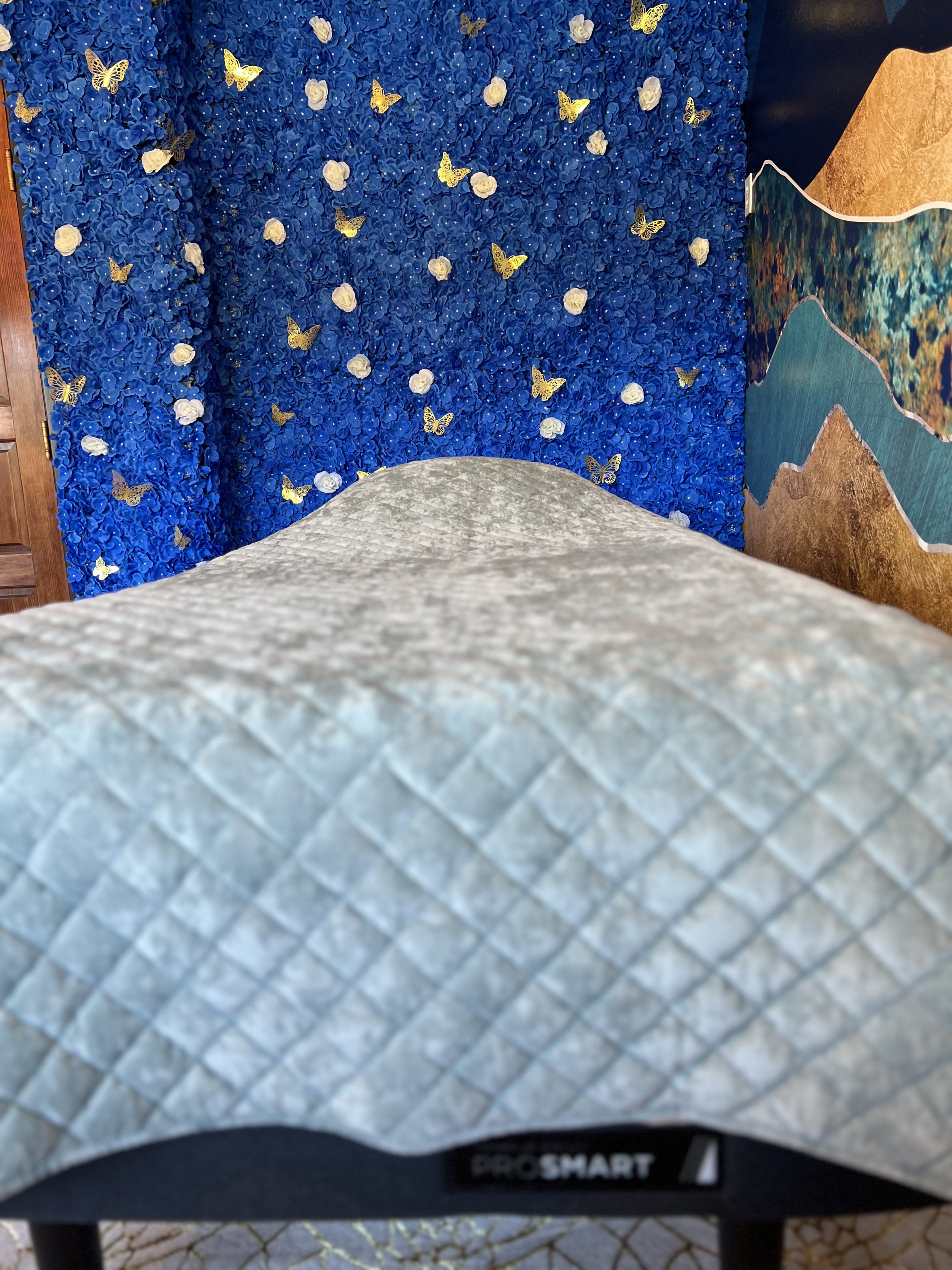 View from the foot of a massage table with a grey quilted cover, facing a blue wall decorated with white roses and gold butterflies, and part of a colorful art piece to the right.