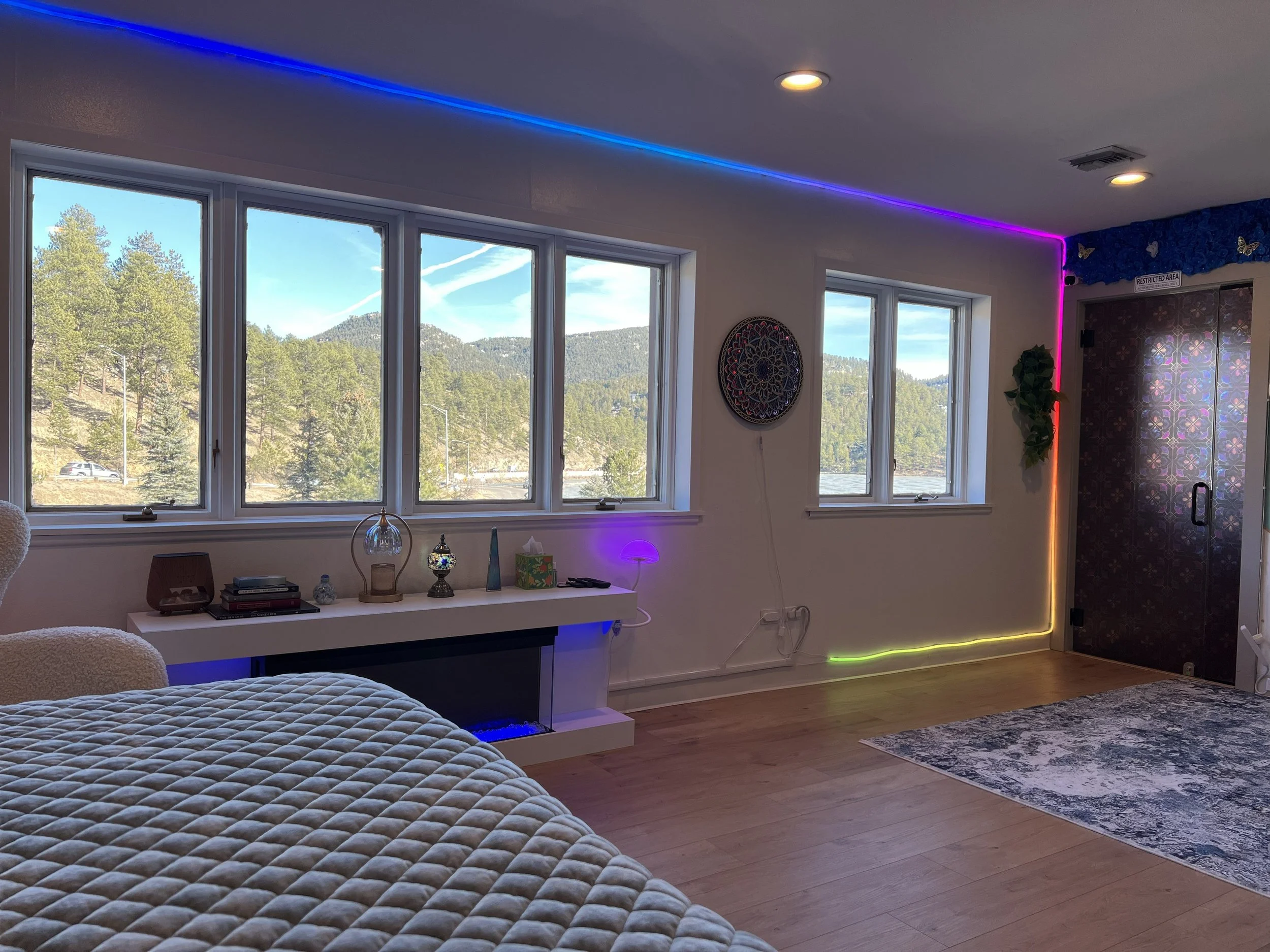 A room with large windows showing a mountain landscape and trees outside, decorated with colorful LED strip lights along the ceiling and wall edges, a white textured bedspread, a white shelf with decorative items, and a patterned rug on a wooden floo