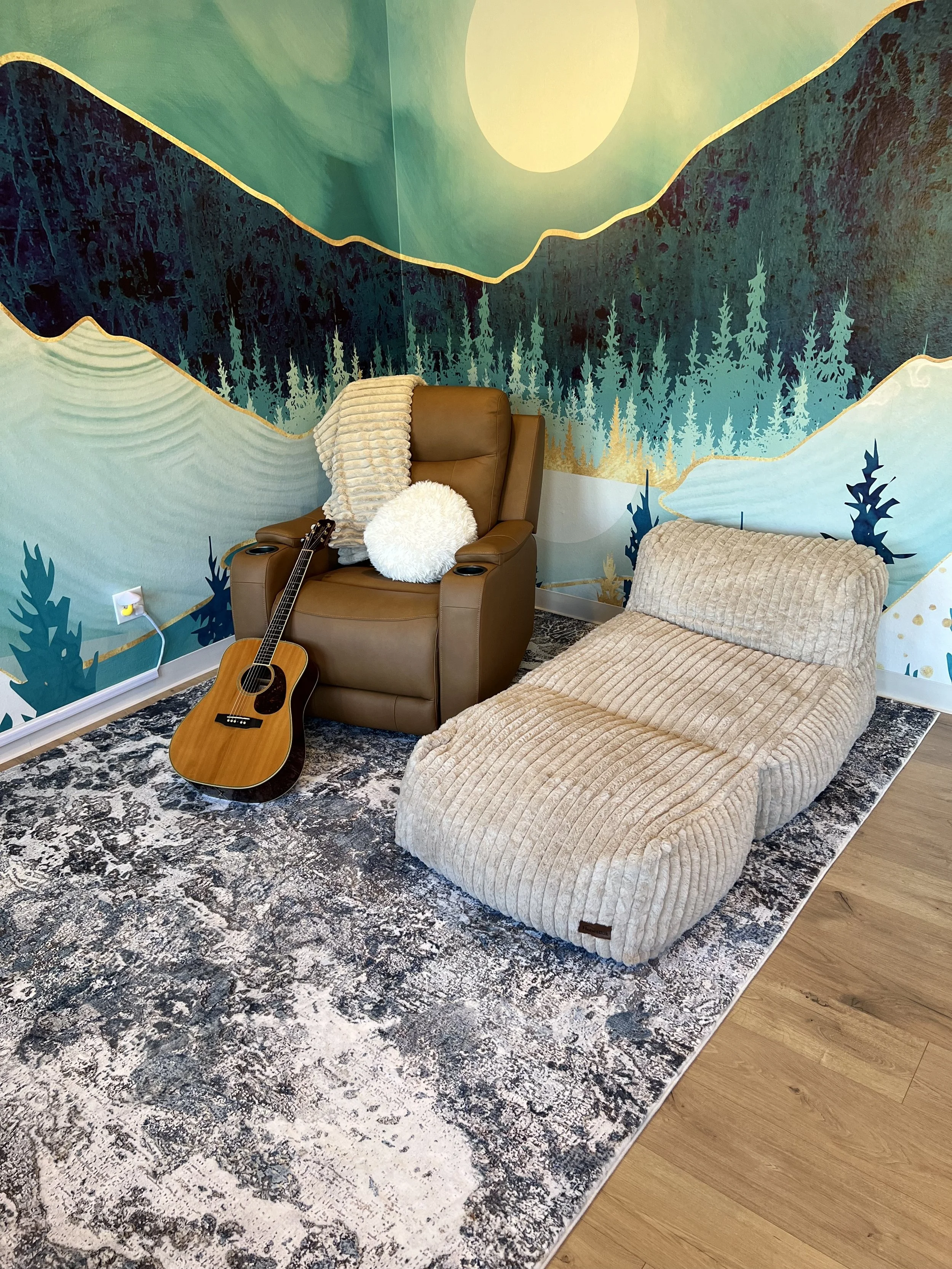Cozy room with a nature-themed wall mural, featuring trees and a large sun or moon. There is a brown recliner chair with a beige throw blanket, a fluffy round pillow, and a guitar leaning against it. Next to the chair is a beige, corduroy lounge chai
