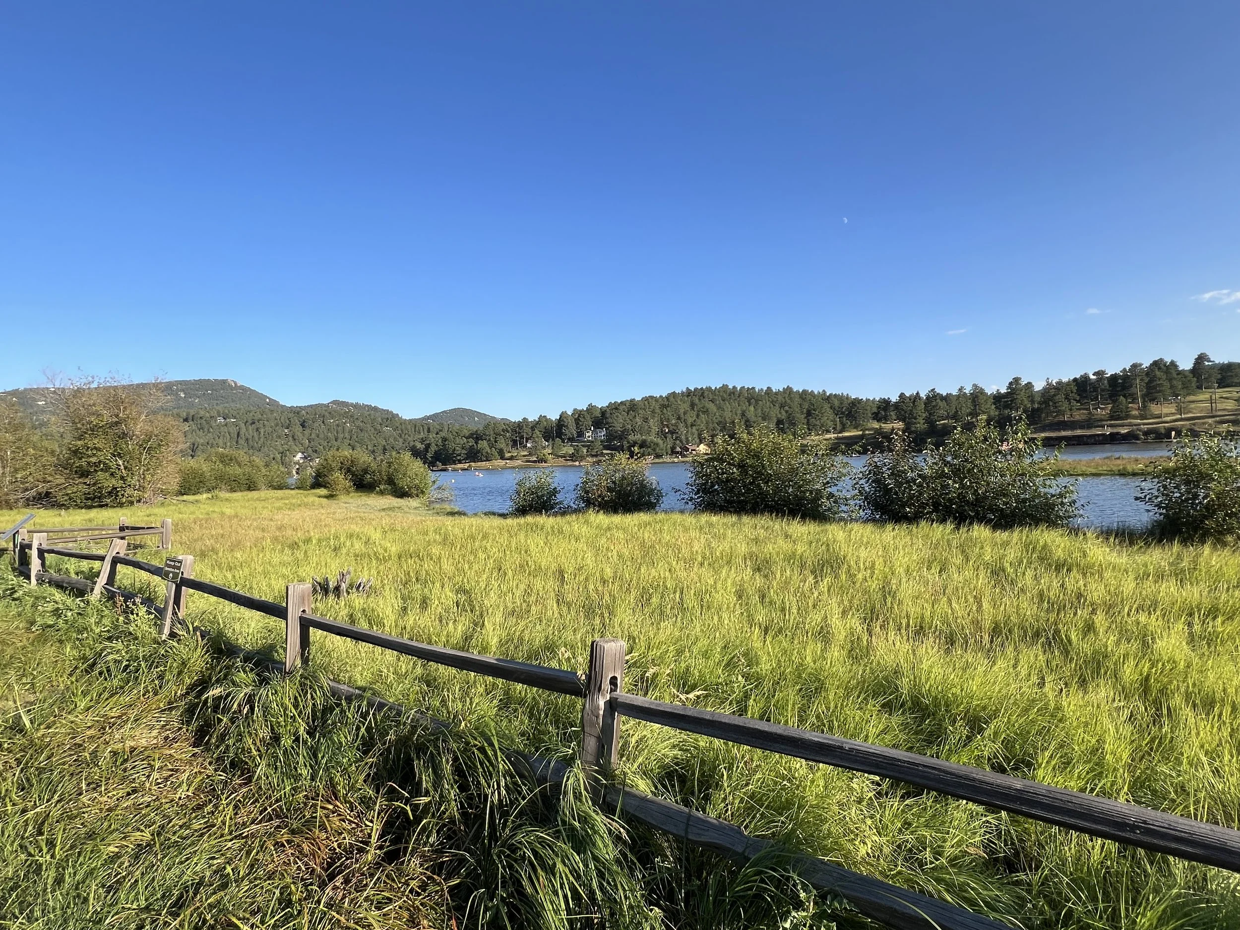 Open grassy field with a wooden fence along the front, a calm lake with trees on the shoreline, hills and mountains in the background, and a clear blue sky above.