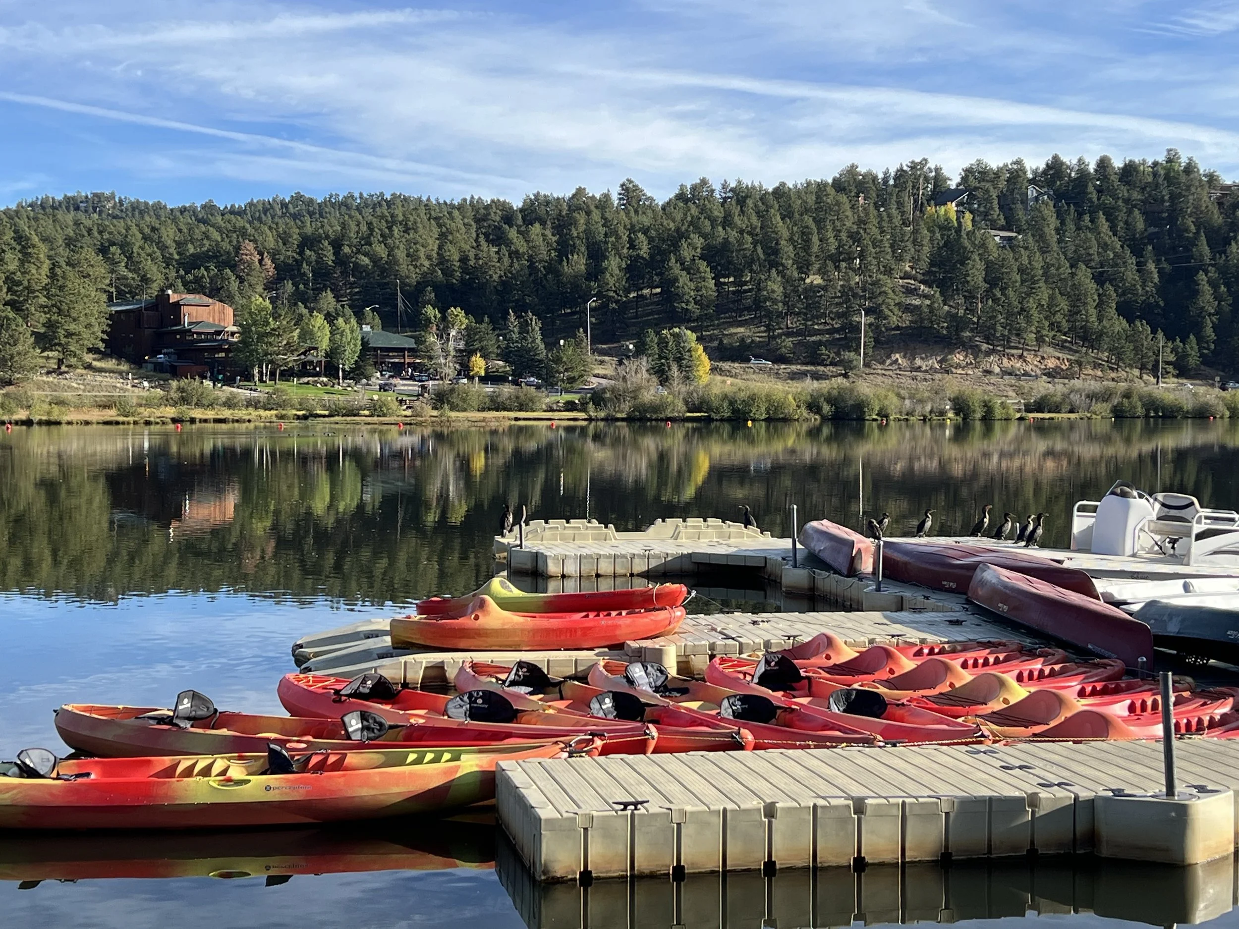 Red and yellow kayaks docked at a marina by a calm lake, with a forested hill and houses in the background under a blue sky with streaks of clouds.