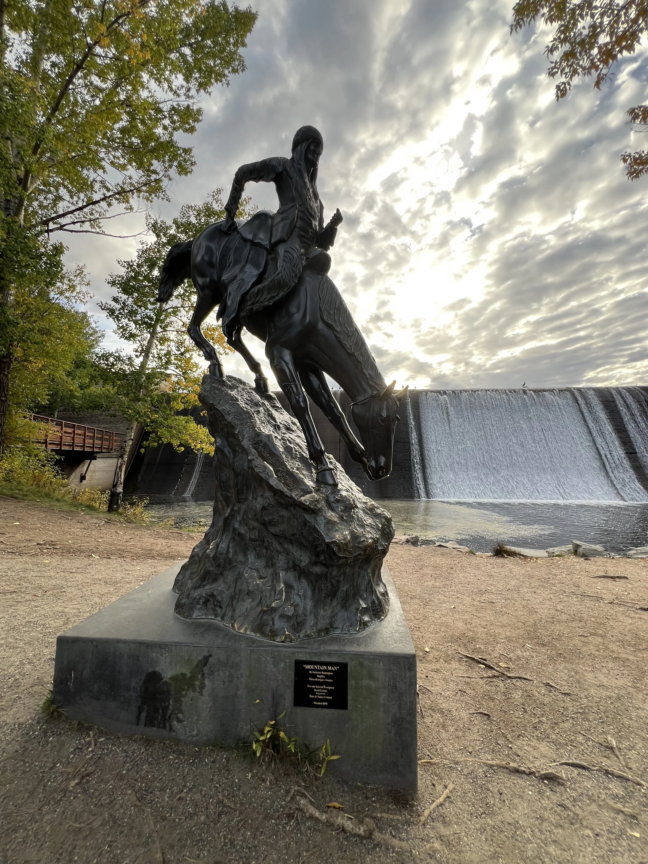 Bronze statue of a cowboy riding a bucking horse against a cloudy sky and a waterfall in the background.