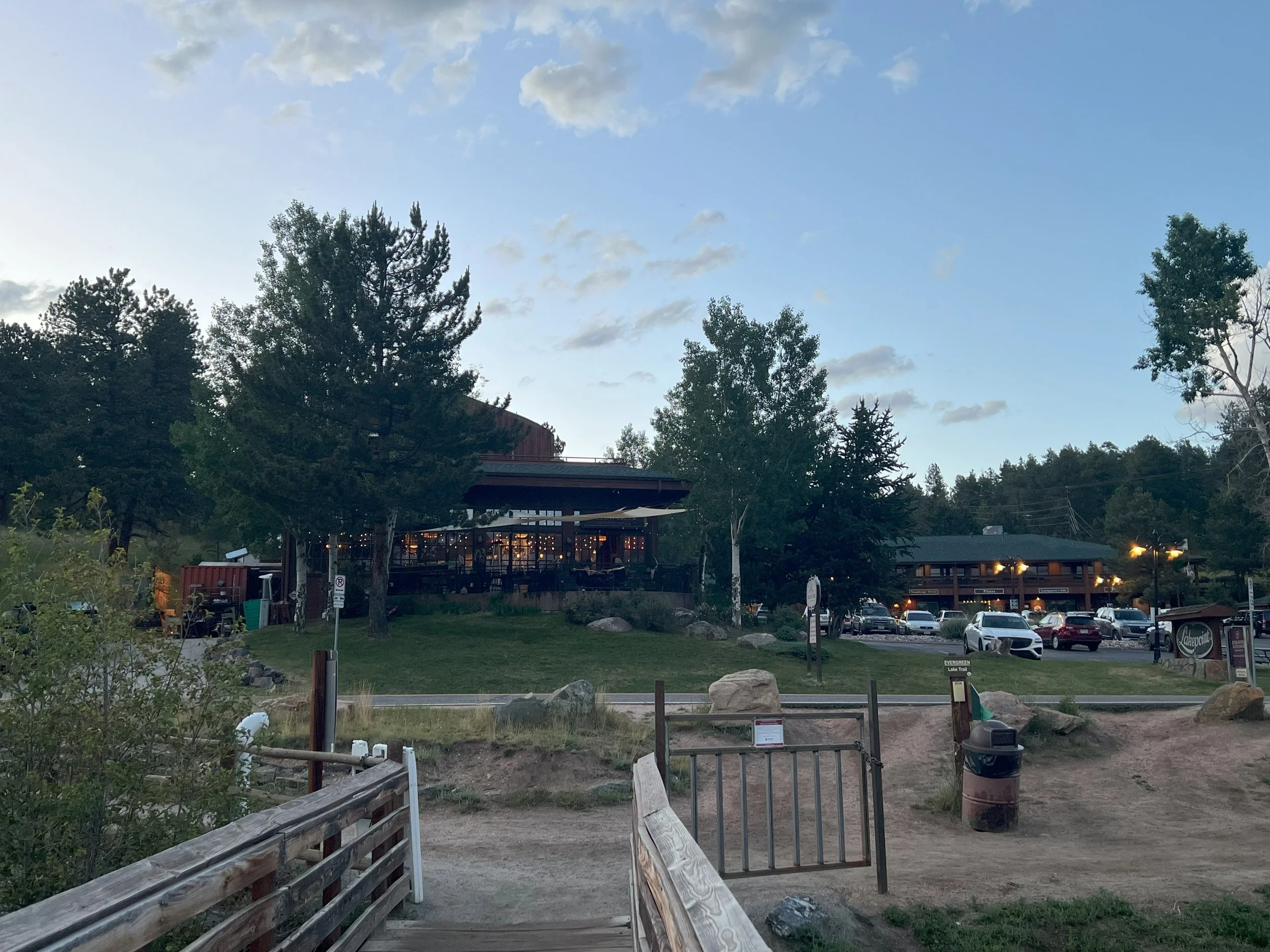 A scenic outdoor view of a parking lot and a restaurant or lodge built among trees, with cars parked and a grassy area with rocks in the foreground.