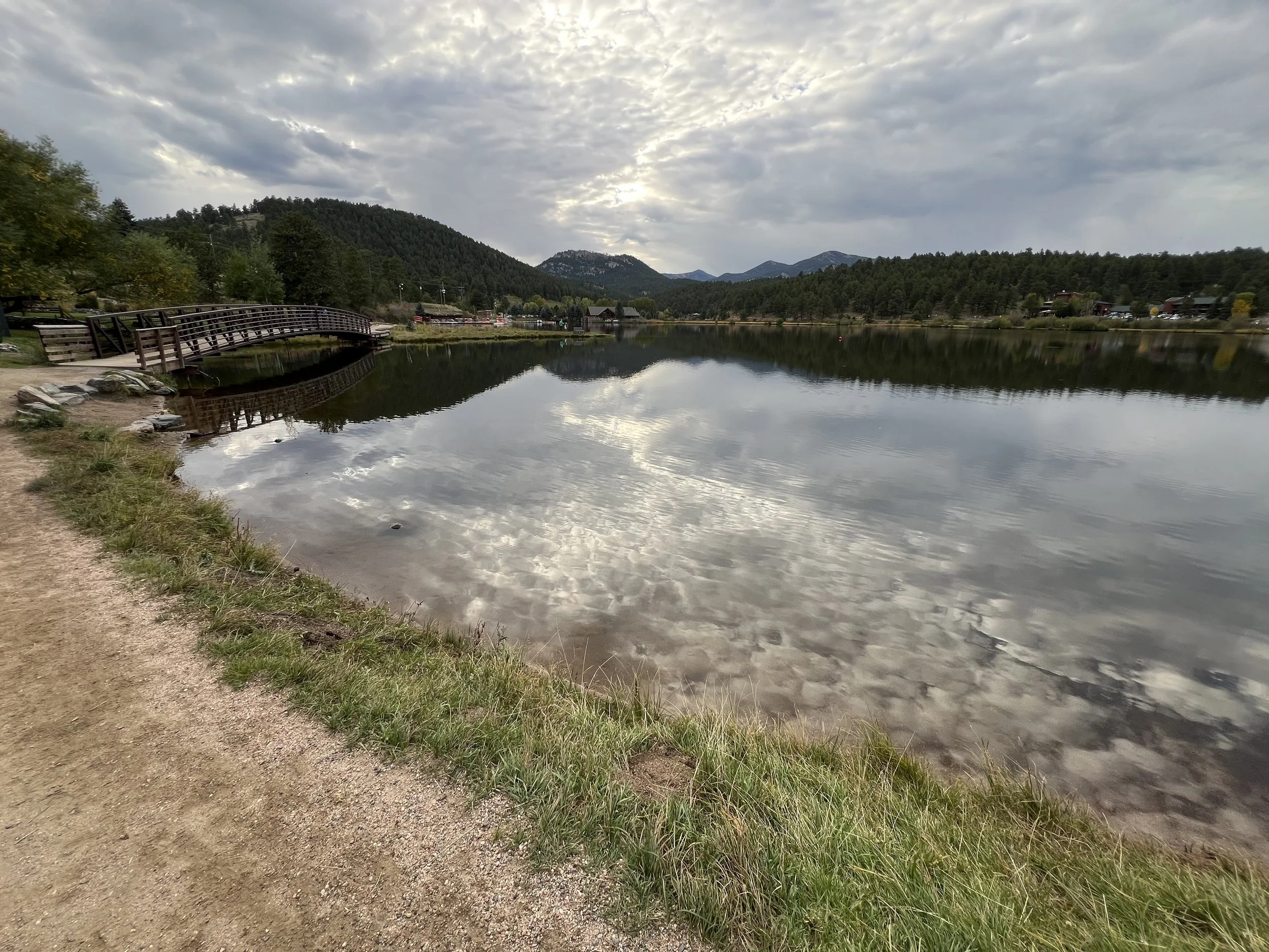 A serene lake with calm water reflecting the cloudy sky above, a wooden bridge on the left, green trees and mountains in the background, and a dirt path along the grassy shoreline.