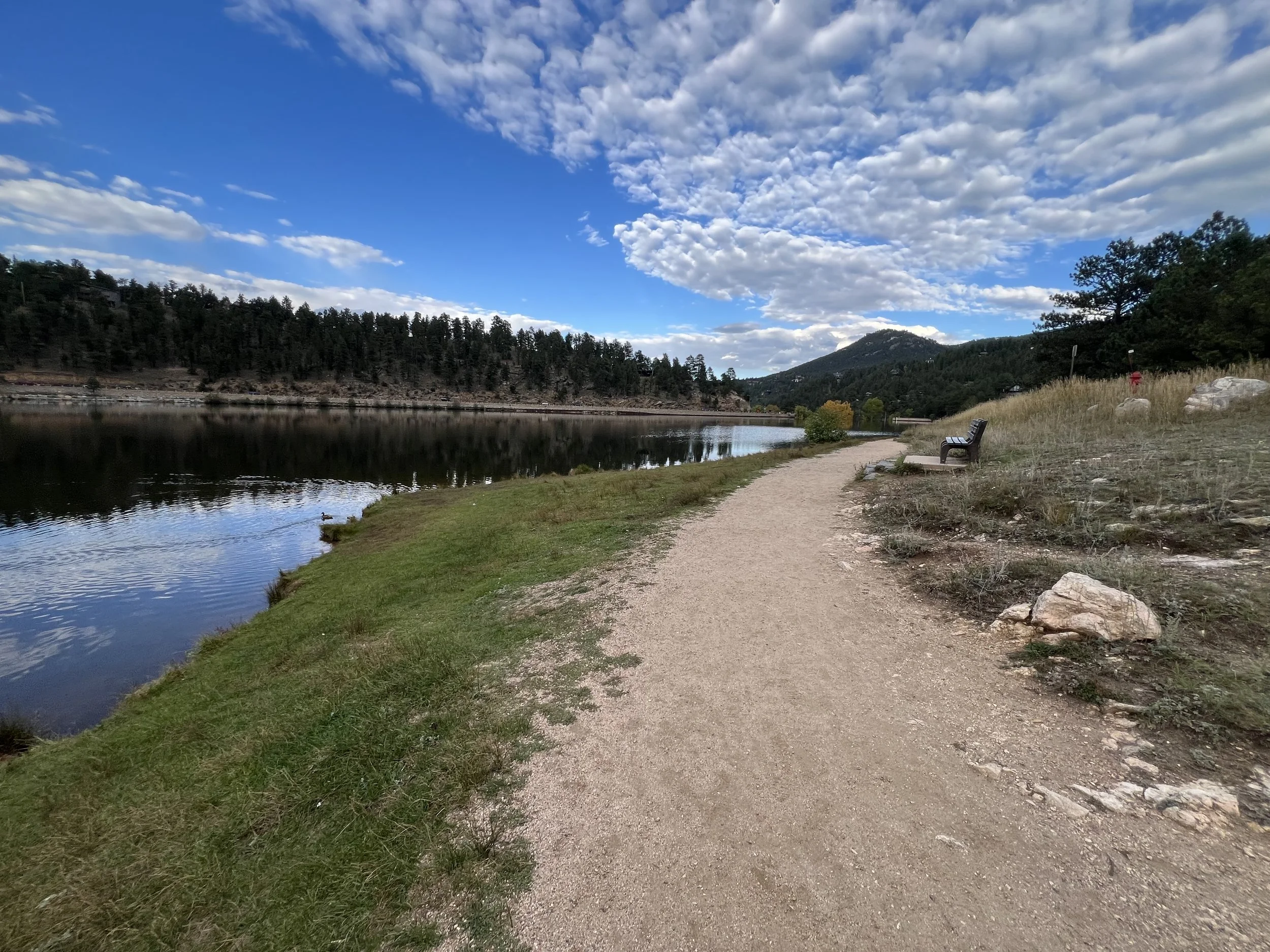 Scenic view of a walking trail along a calm river with trees, hills, and a partly cloudy sky in the background.