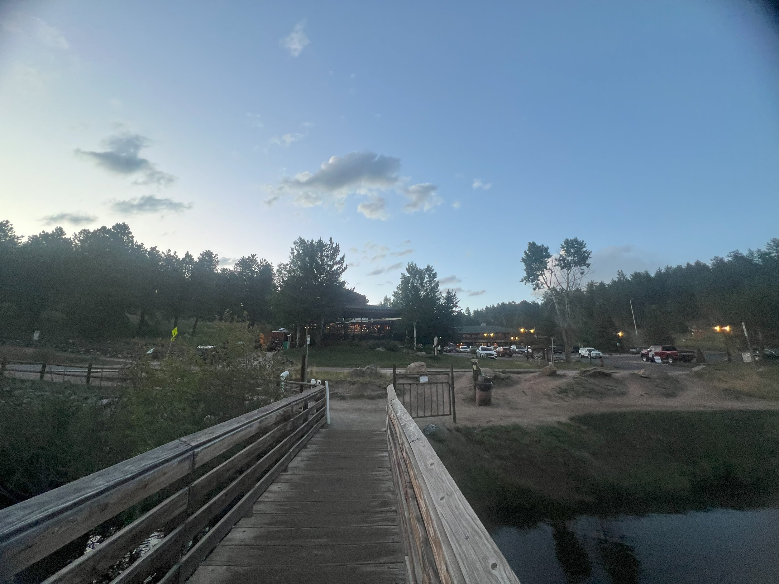 Wooden bridge over a small water body, with trees, parking lot, and building in the distance during evening.
