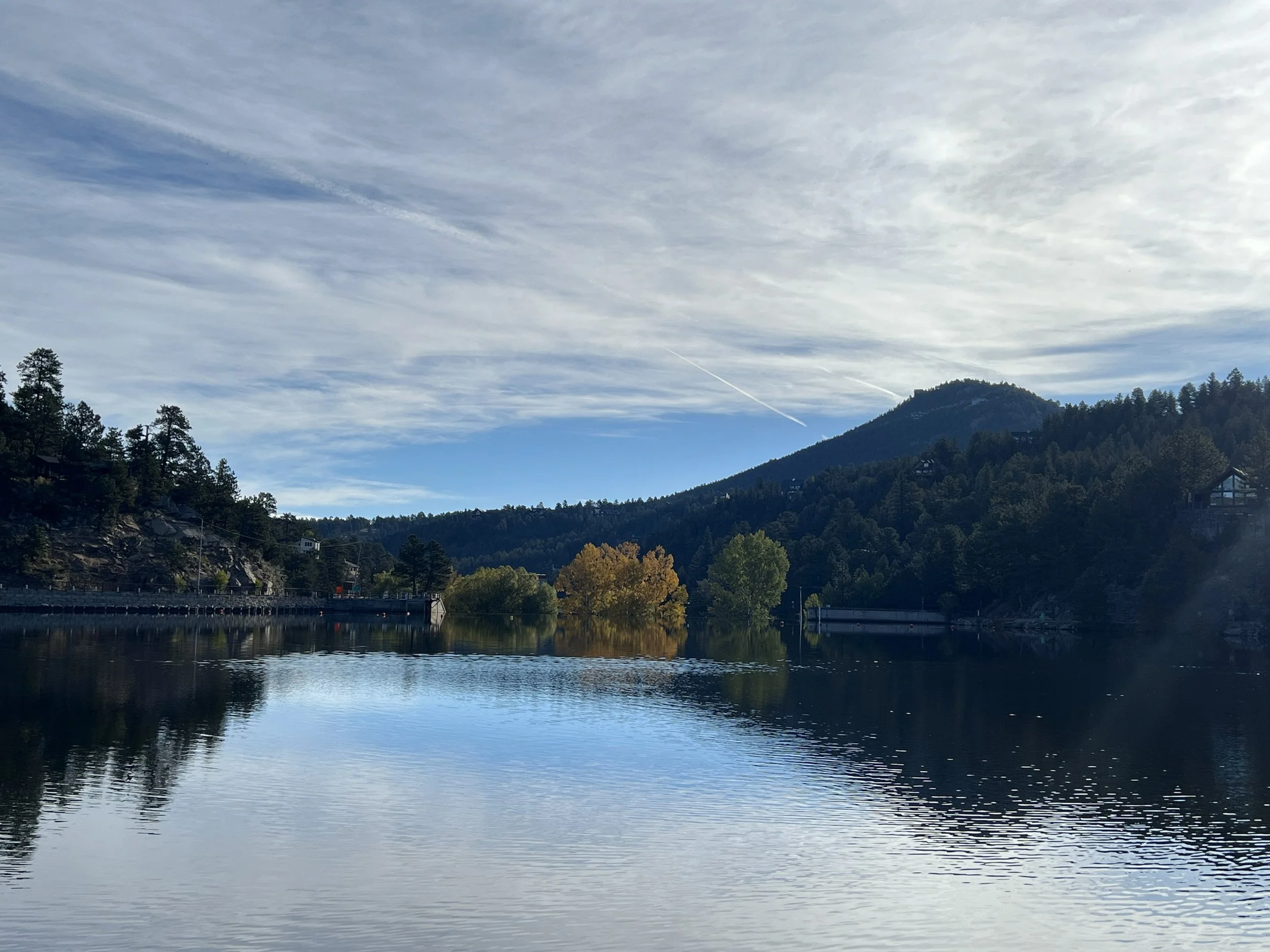 A serene lake surrounded by trees and hills, with a partly cloudy sky overhead and the water reflecting the landscape.
