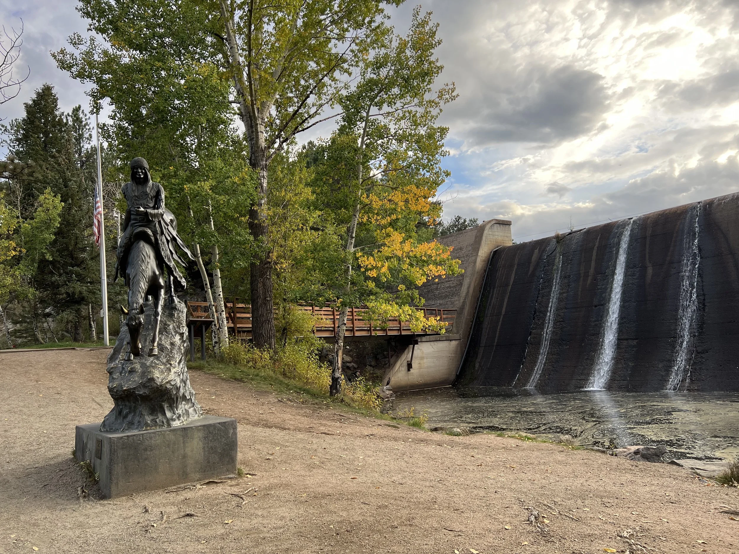 A statue of a woman riding a horse on a rocky base, with an American flag nearby, trees with green and yellow leaves, a dam with water cascading over it, and a cloudy sky in the background.