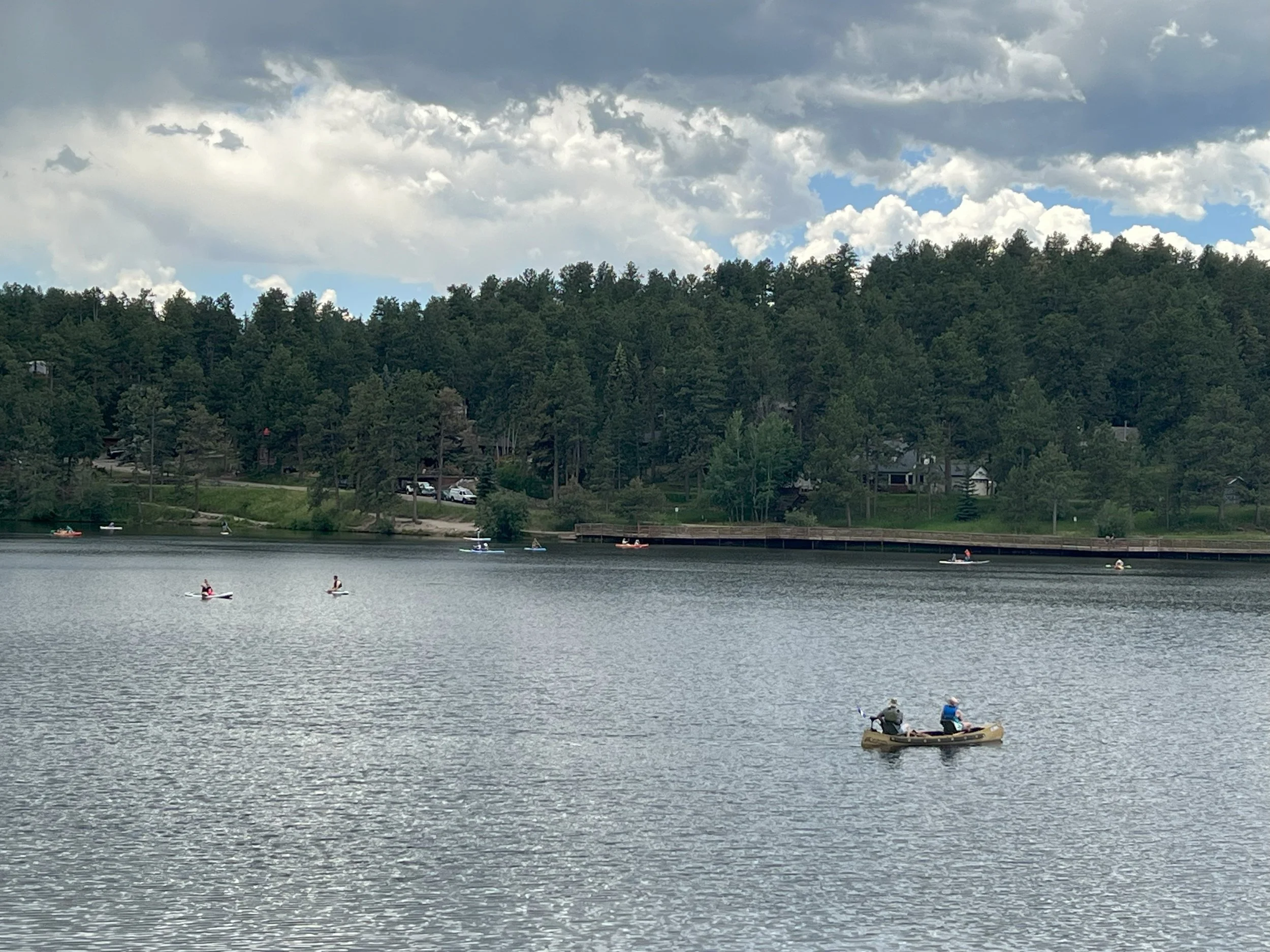 People kayaking and paddleboarding on a calm lake surrounded by trees and a hillside with houses.