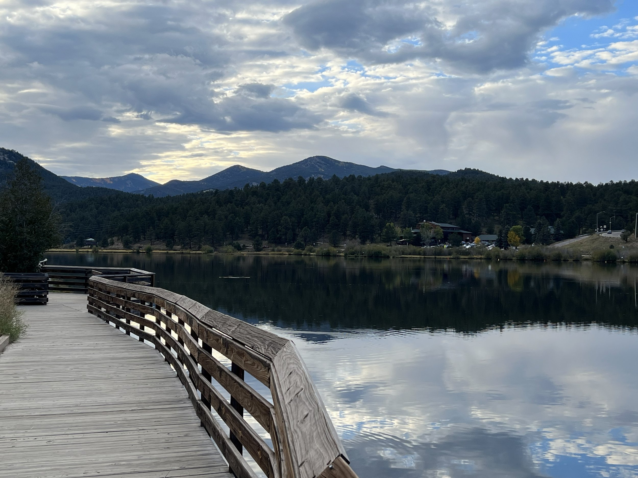 A wooden dock extends over a calm body of water, surrounded by a forested landscape with mountains in the background and a partly cloudy sky overhead.