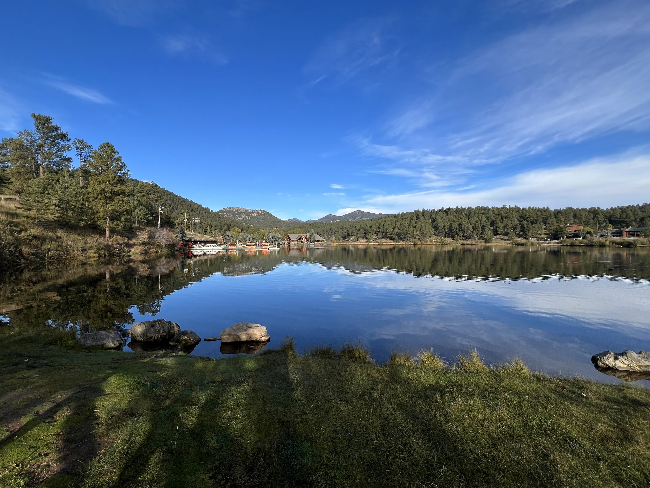 Scenic view of a calm lake with reflections of trees and mountains under a partly cloudy blue sky.