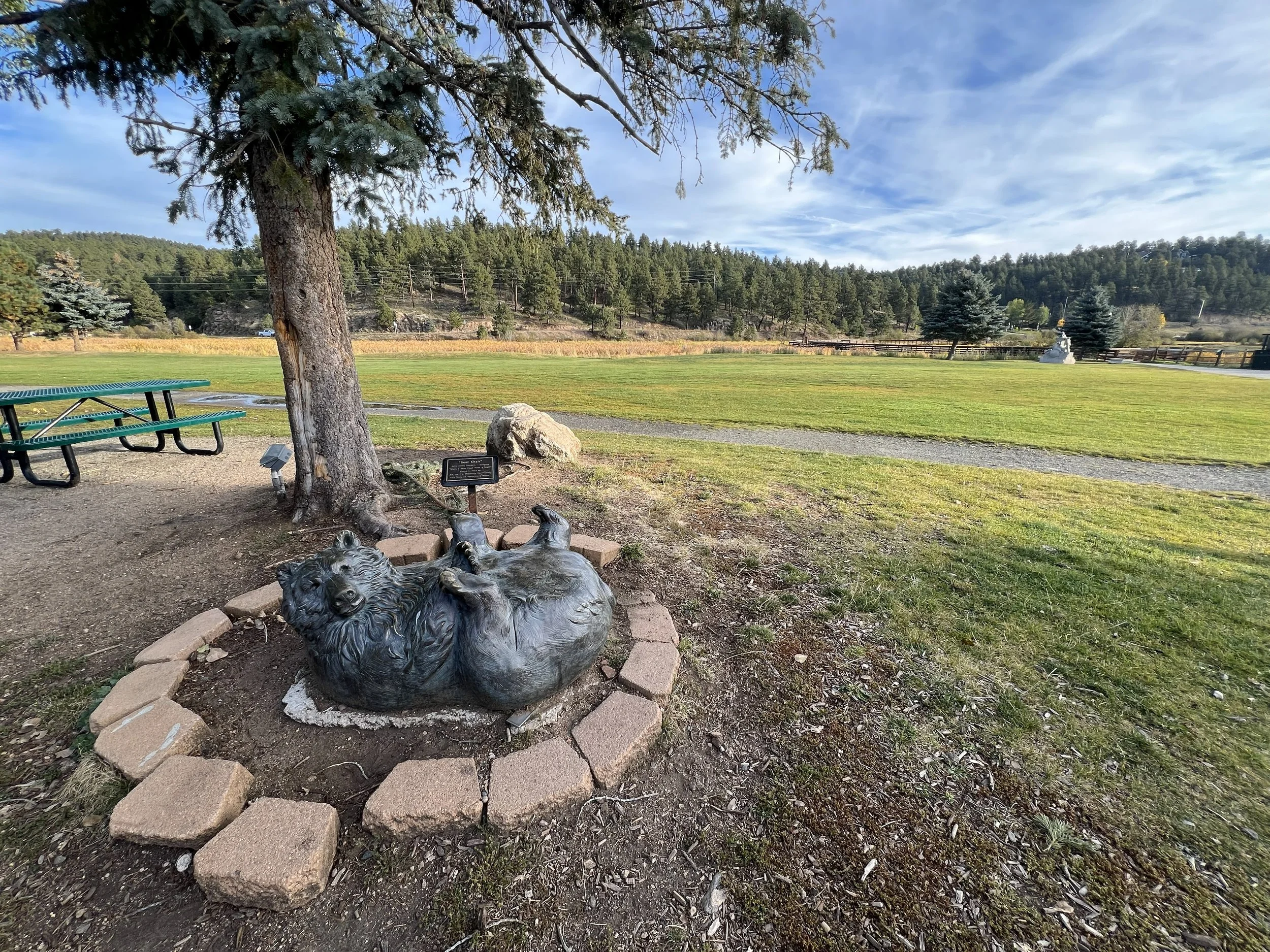 A bronze sculpture of a bear lying on its back, surrounded by a brick border, located under a large tree in a park with open grassy fields and distant trees.