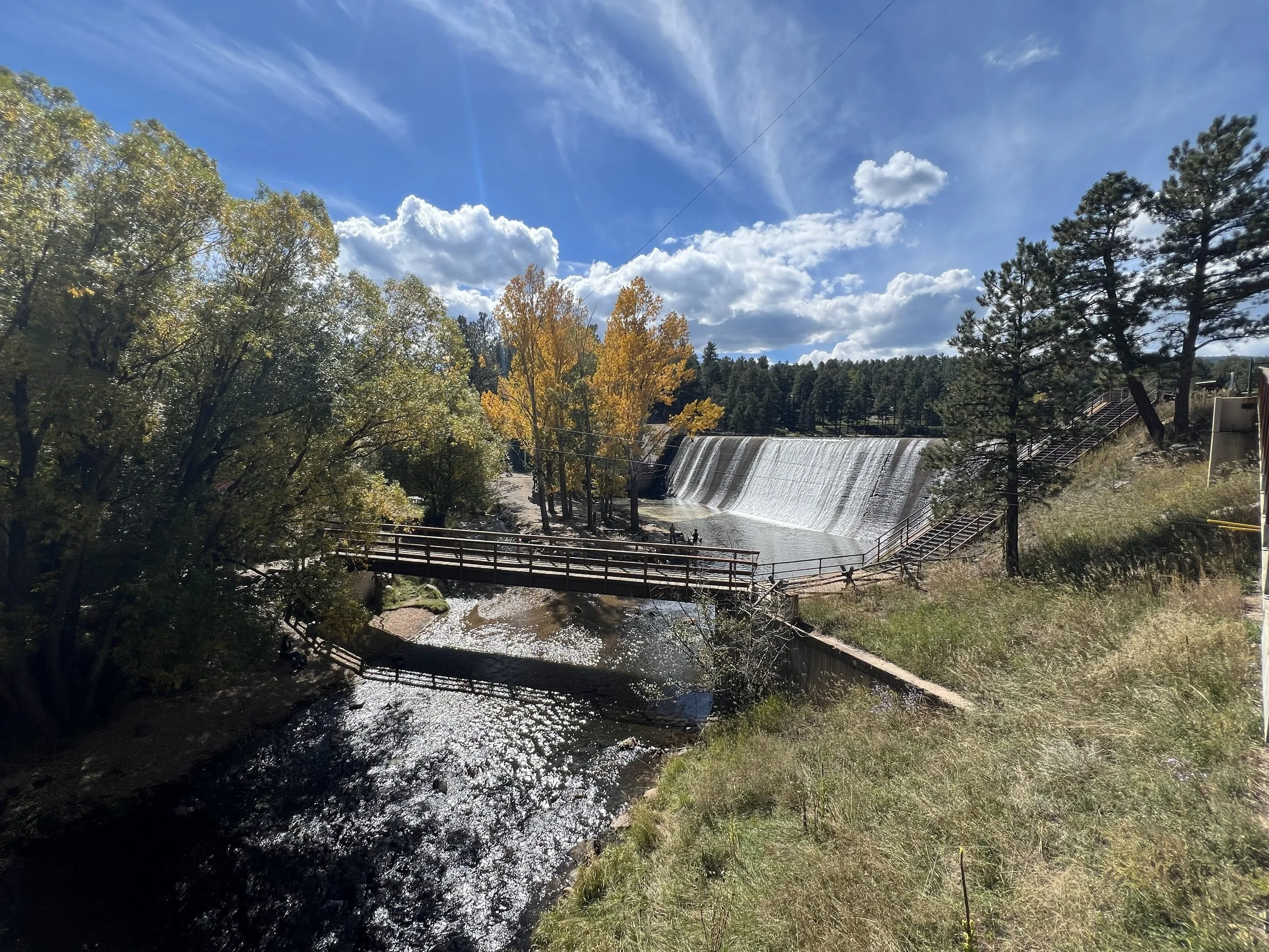 A scenic view of a waterfall with a small bridge and walking path, surrounded by trees with fall foliage, under a partly cloudy sky.