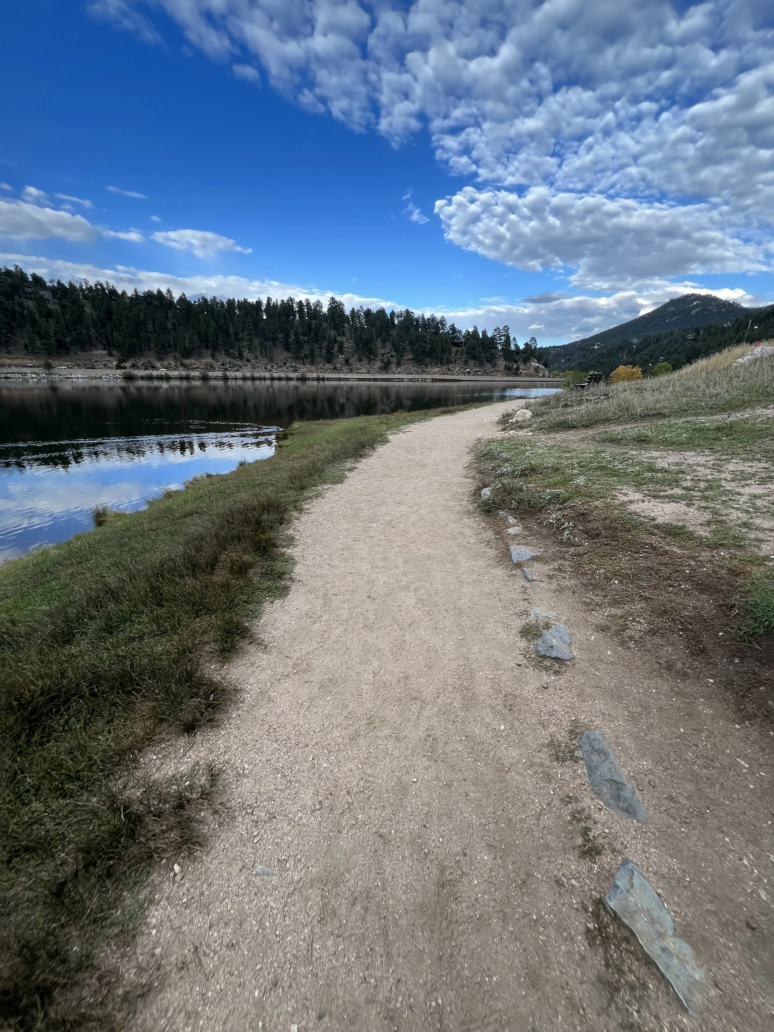 A dirt trail runs alongside a body of water with trees on the far side, surrounded by mountains and a partly cloudy blue sky.