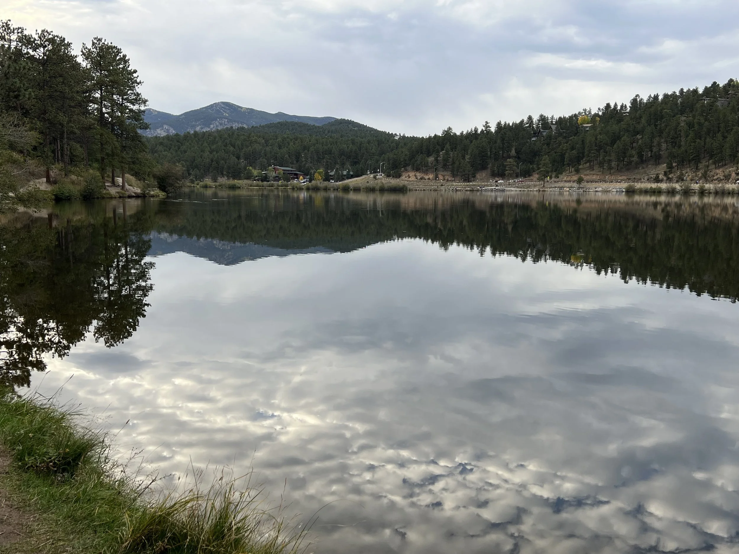 Reflection of cloudy sky, trees, and mountains on a calm lake during daytime.
