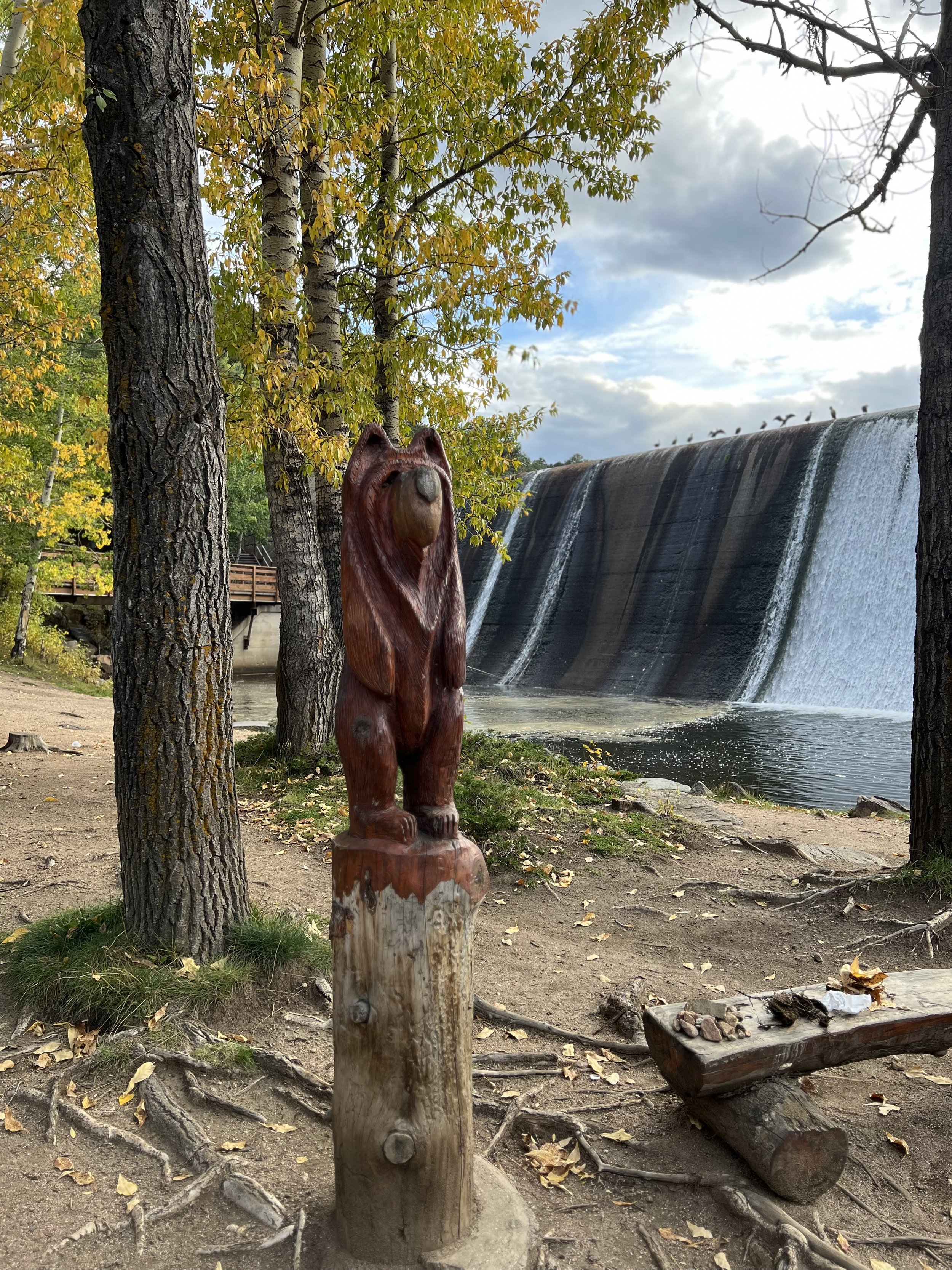 A wooden bear sculpture standing on a tree stump in a wooded area with a waterfall in the background, trees with green and yellow leaves, and a partly cloudy sky.