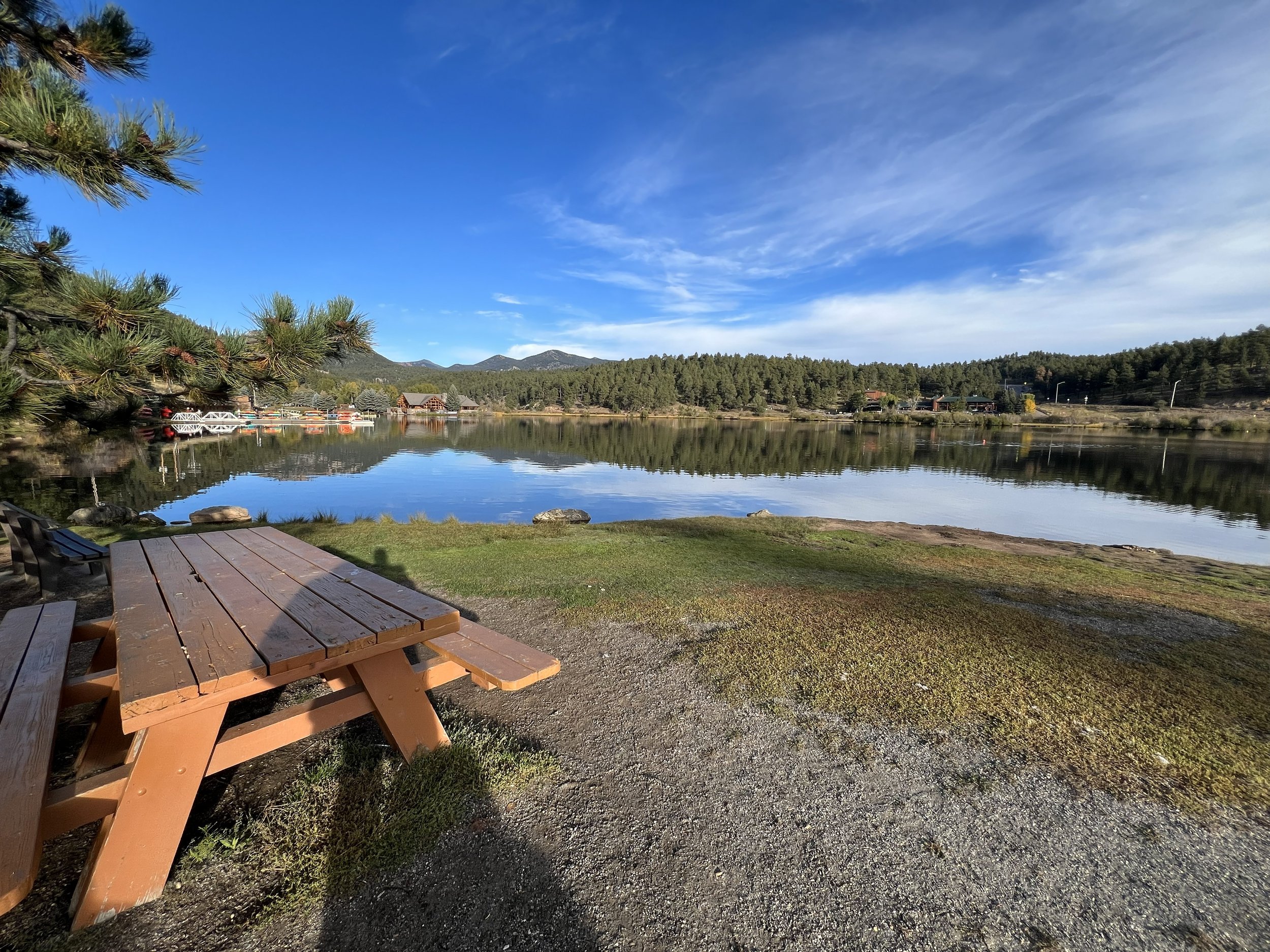 A lakeside scene with a wooden picnic table and bench in the foreground, calm water reflecting the blue sky and clouds, surrounded by greenery and trees, with houses and hills in the background.