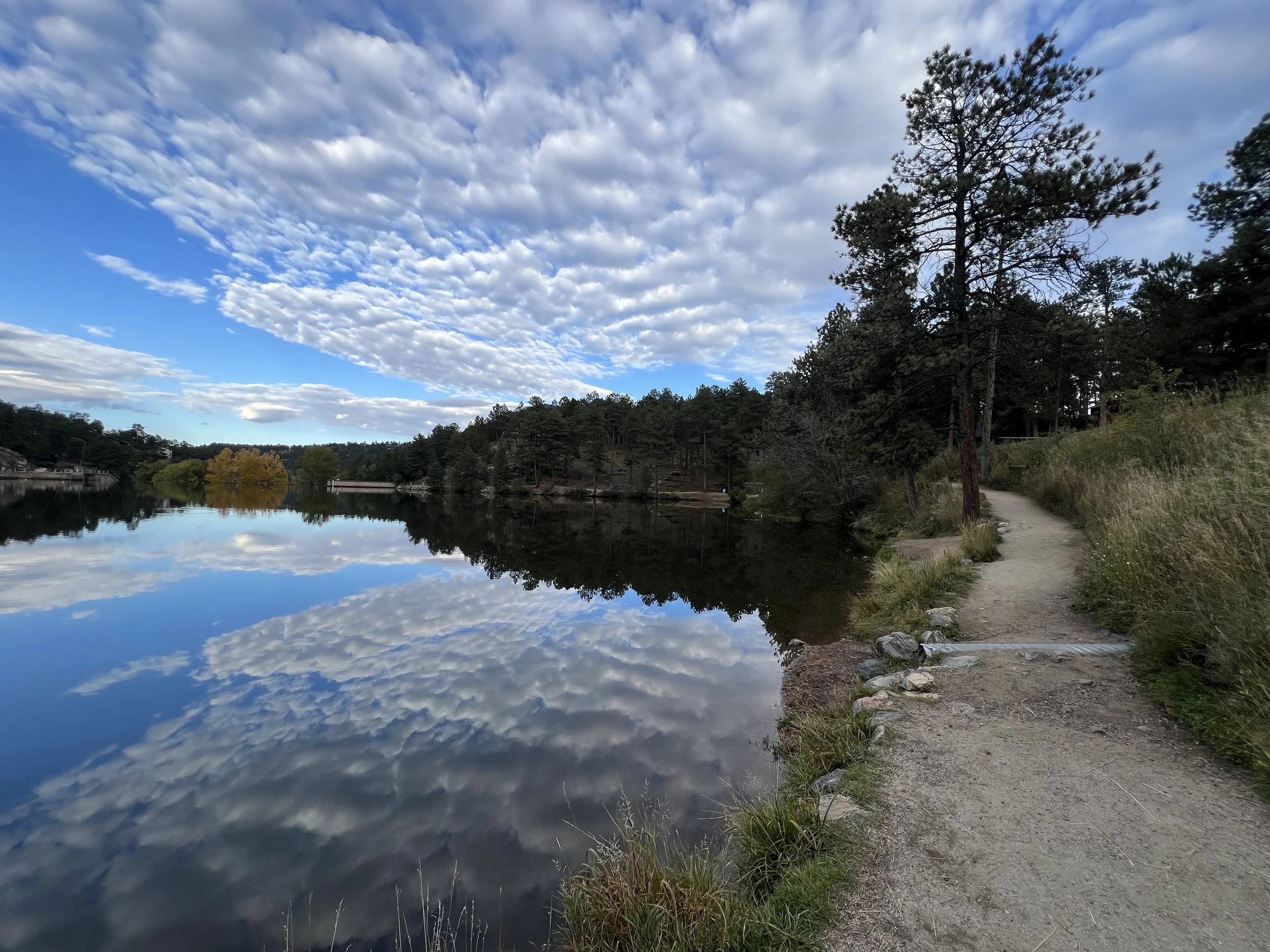 A peaceful lakeside scene with a dirt trail on the right, tall trees along the shore, and a partly cloudy sky reflected in the calm water.