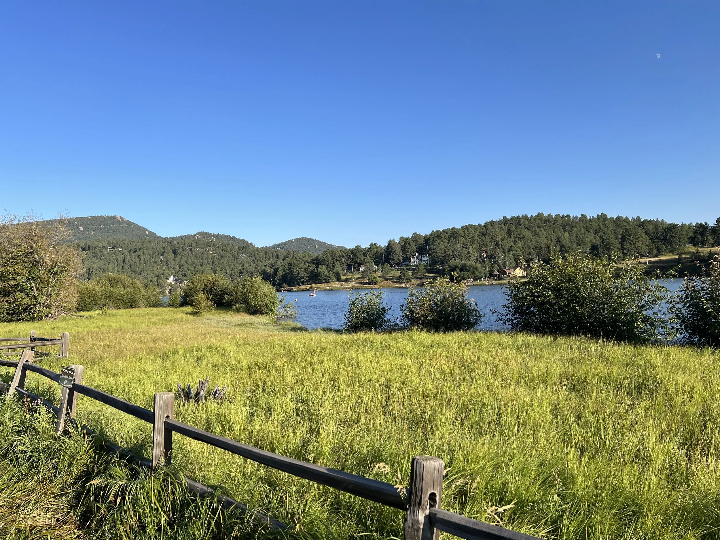 Scenic view of a grassy field with a wooden fence, a lake, and forested hills in the background on a clear day with blue sky and visible moon.