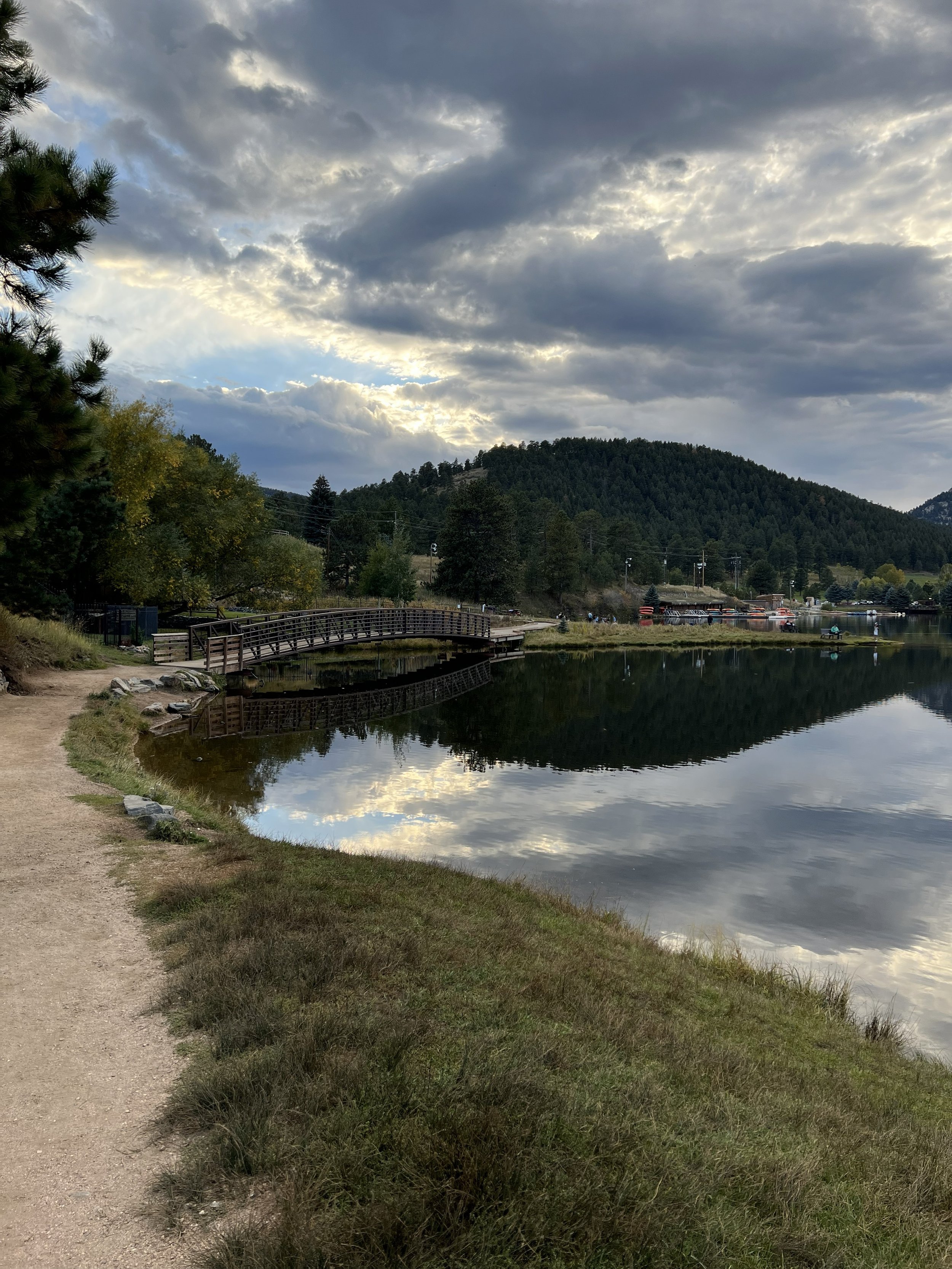 A lakeside scene with a dirt walking path on the left, grassy shoreline, calm water reflecting the cloudy sky, trees, and a small bridge crossing the water. Mountains and a few small boats are visible in the distance.