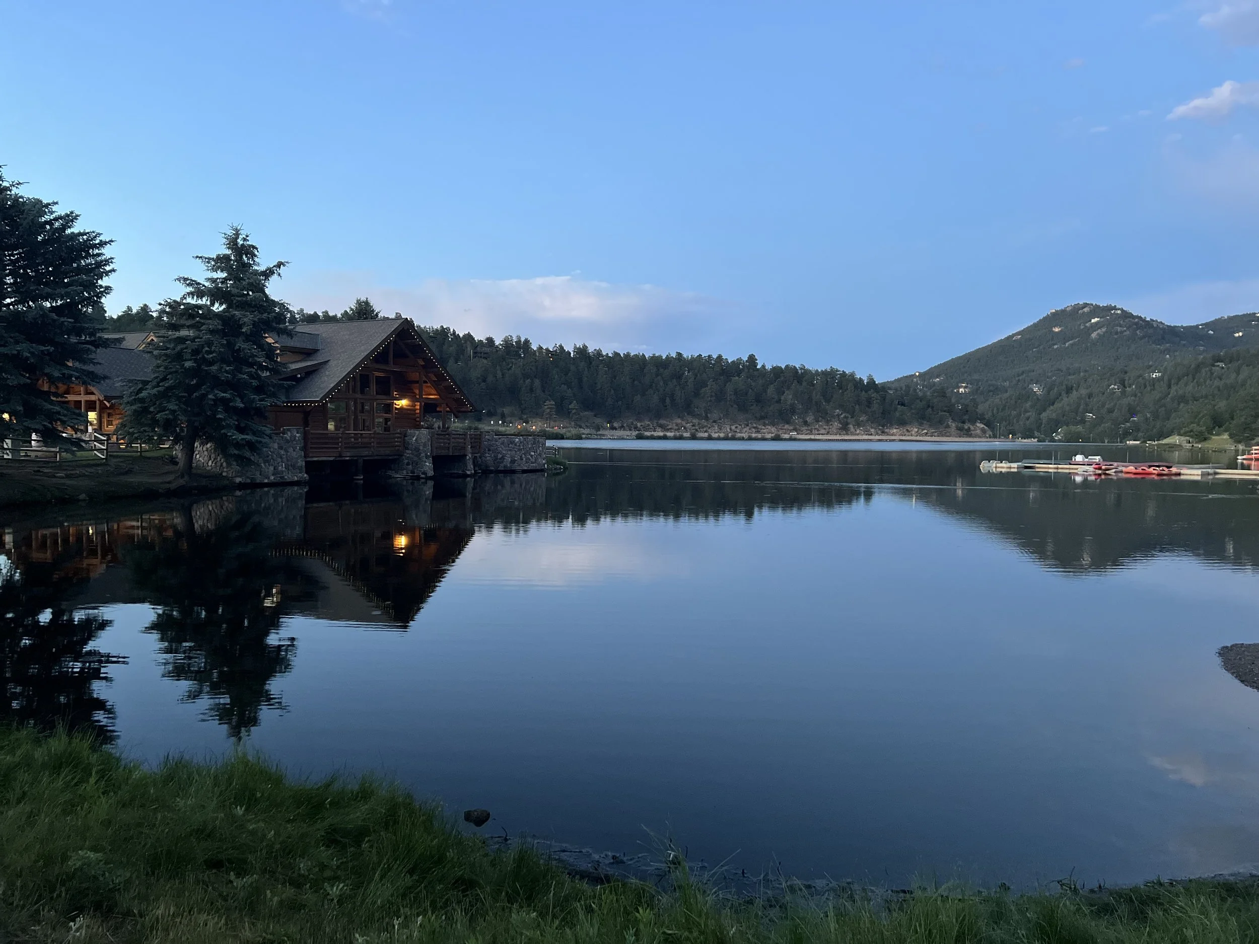 A peaceful lake scene with a wooden lodge and pine trees on the shore, reflecting in the calm water, with mountains in the background and a clear blue sky.