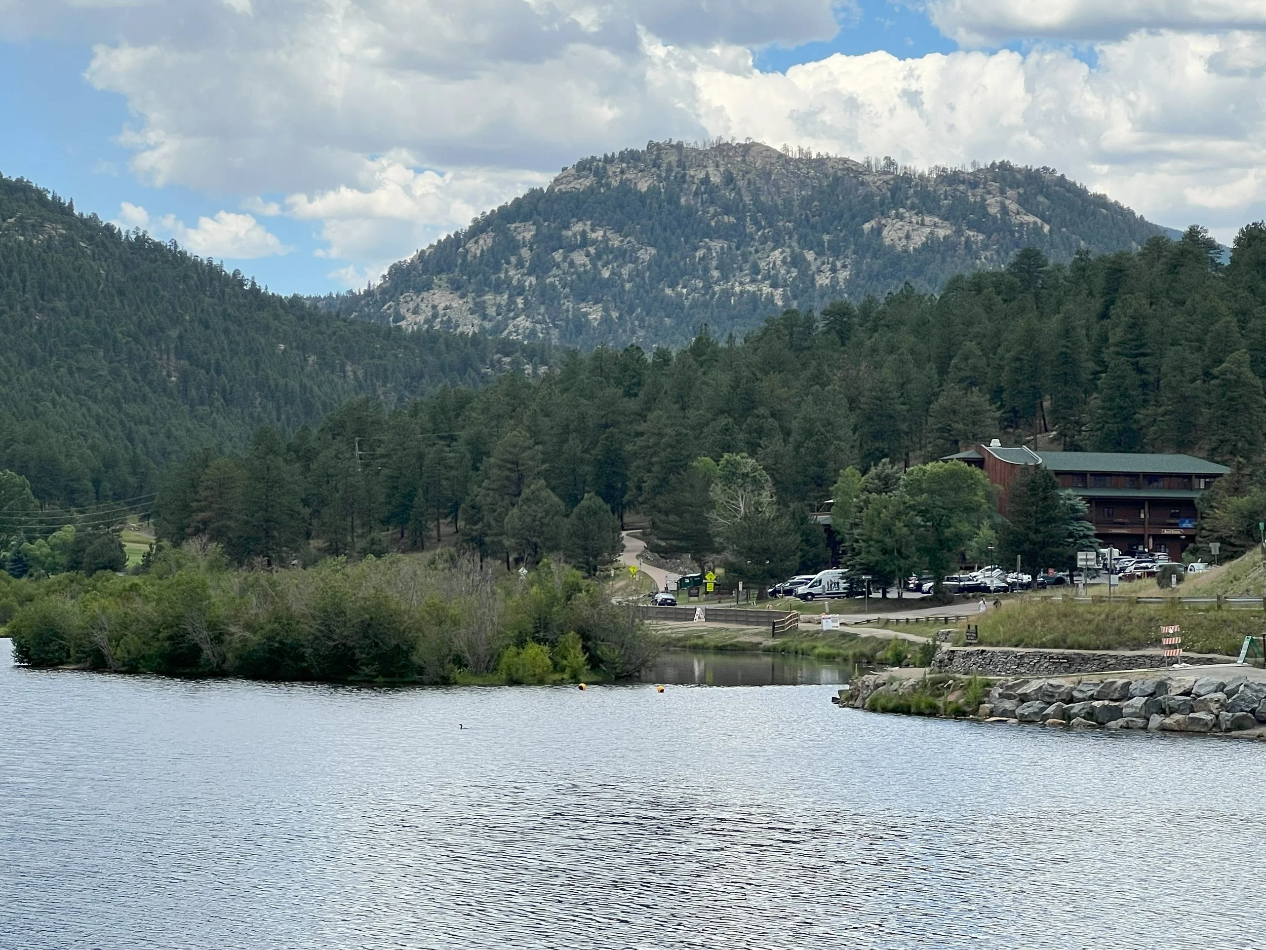 A scenic view of a lake with a rippled surface, surrounded by lush green trees, with mountains and a partly cloudy sky in the background. A parking lot and a wooden building are visible on the side near the trees.