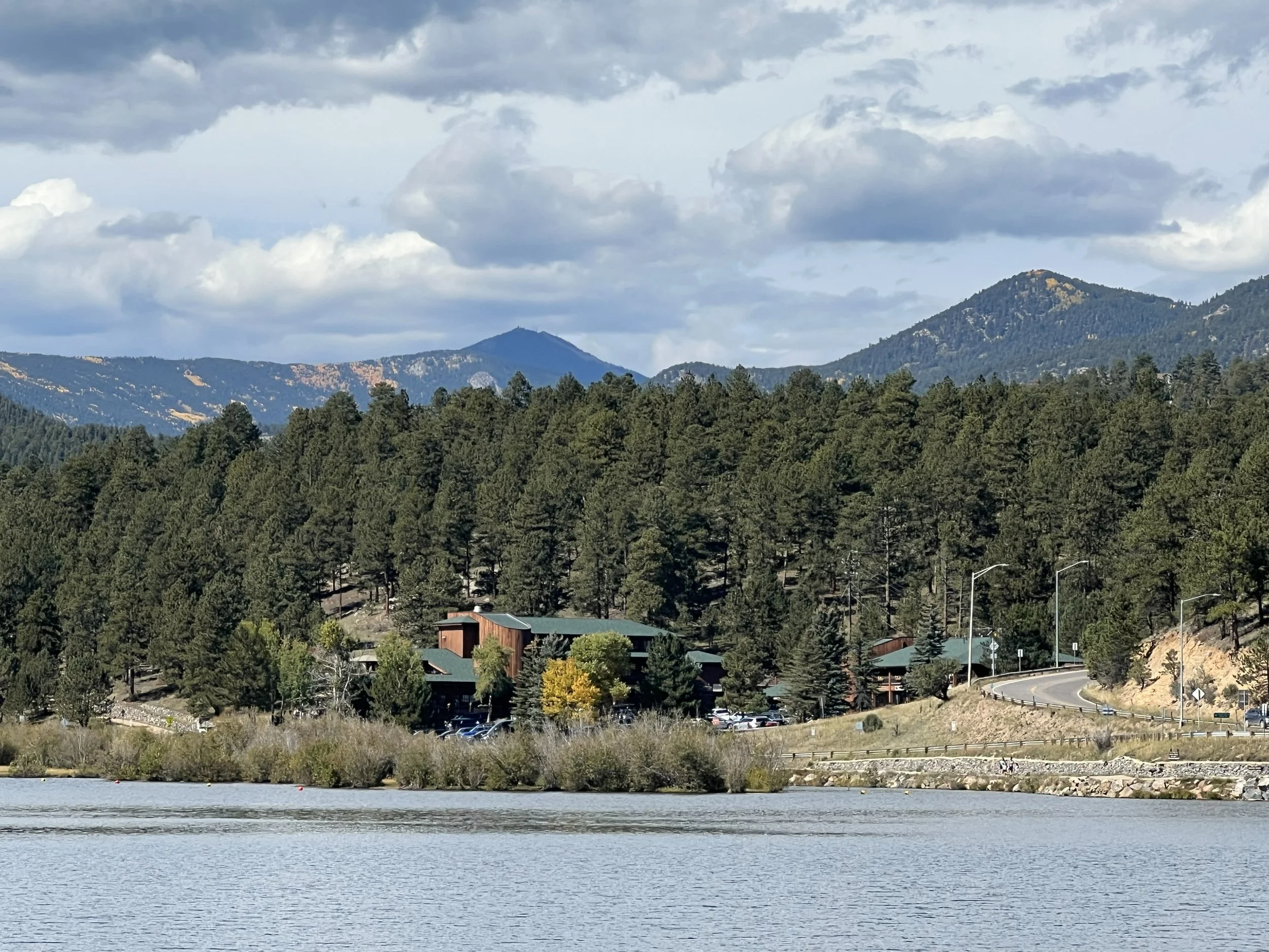 A scenic landscape featuring a calm lake in the foreground, surrounded by dense forest and mountains in the background under a partly cloudy sky.