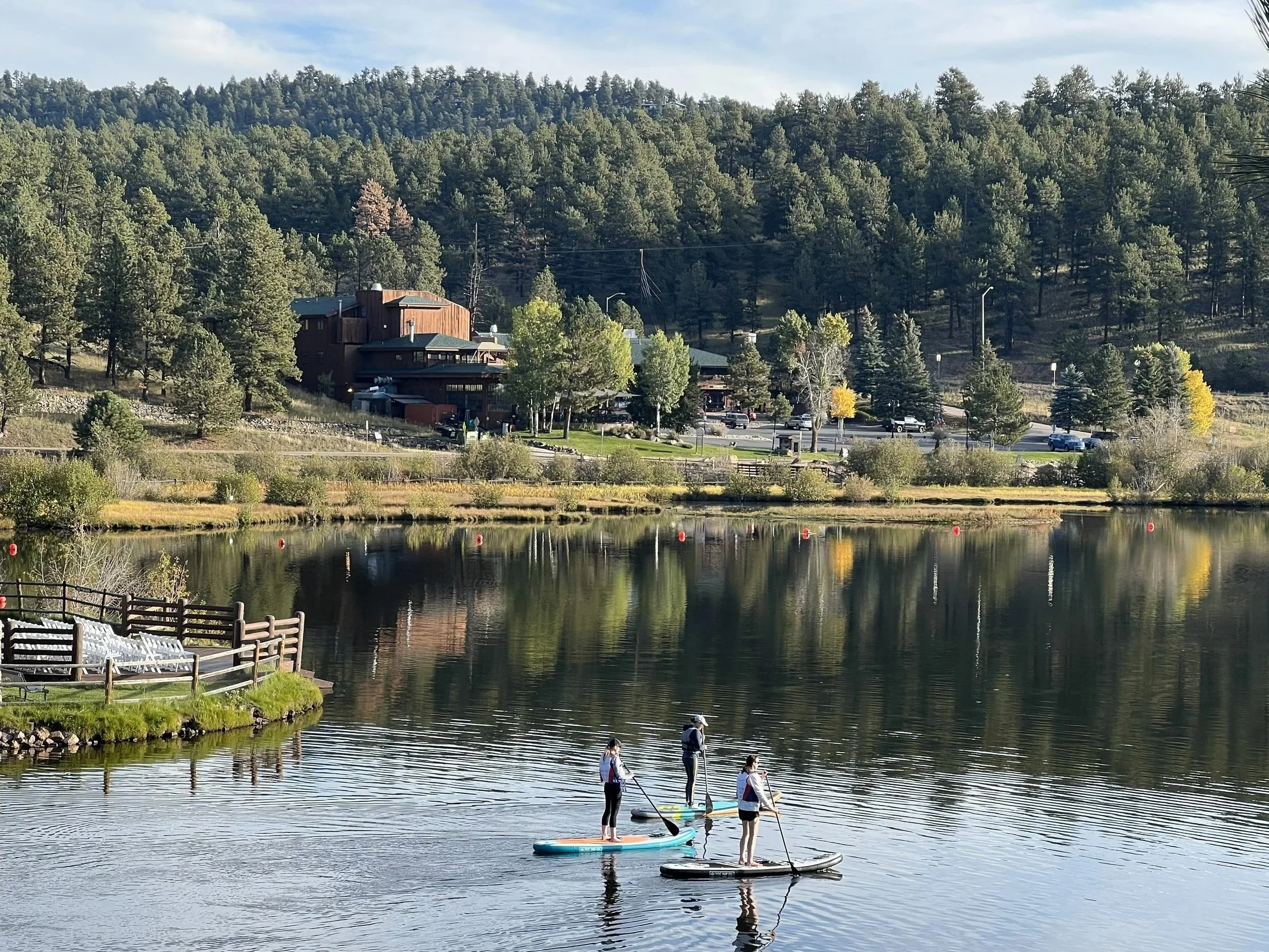 Three people paddleboarding on a calm lake with a wooded hillside and buildings in the background.