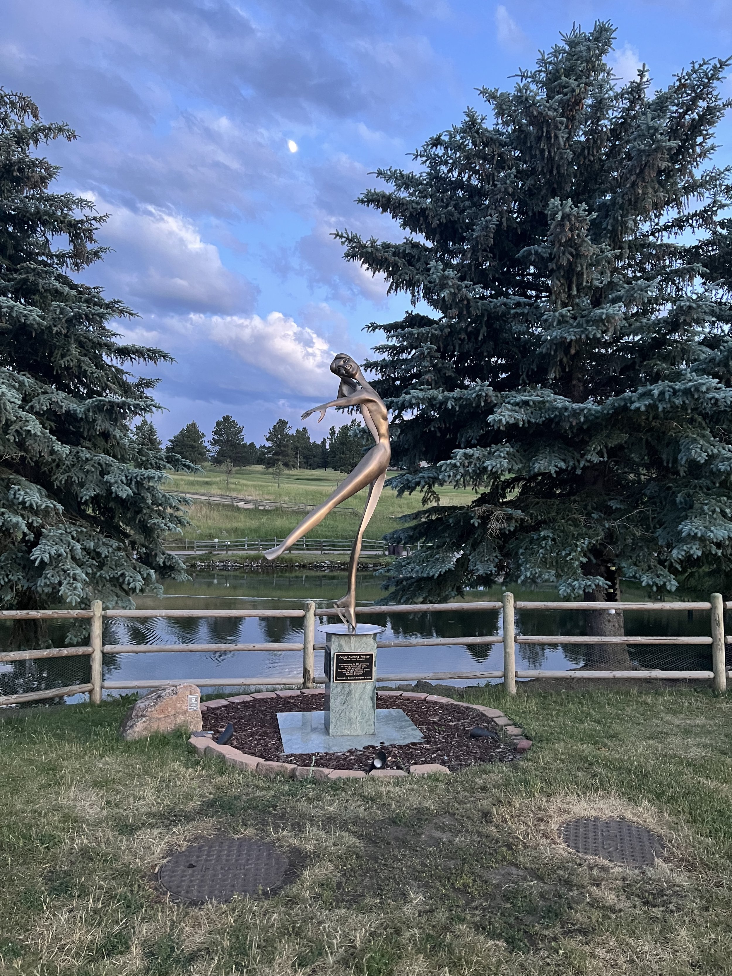 A silver sculpture of a ballerina on a pedestal, surrounded by a small flower bed, with pine trees and a lake in the background under a sky with clouds and the moon.
