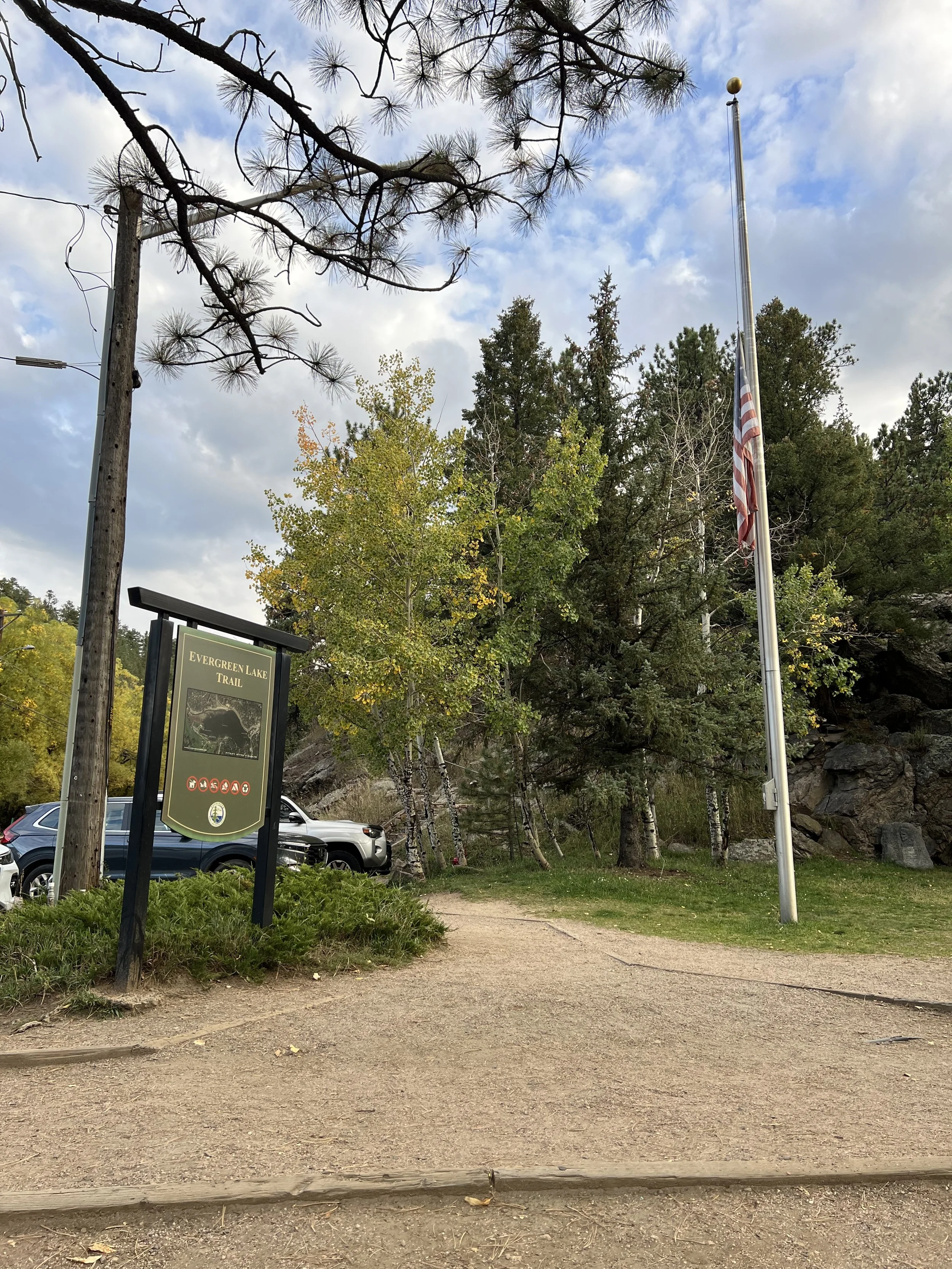 A dirt trailhead marked as 'Evergreen Lake Trail' with parking area, trees, an American flag on a flagpole, and a cloudy sky.