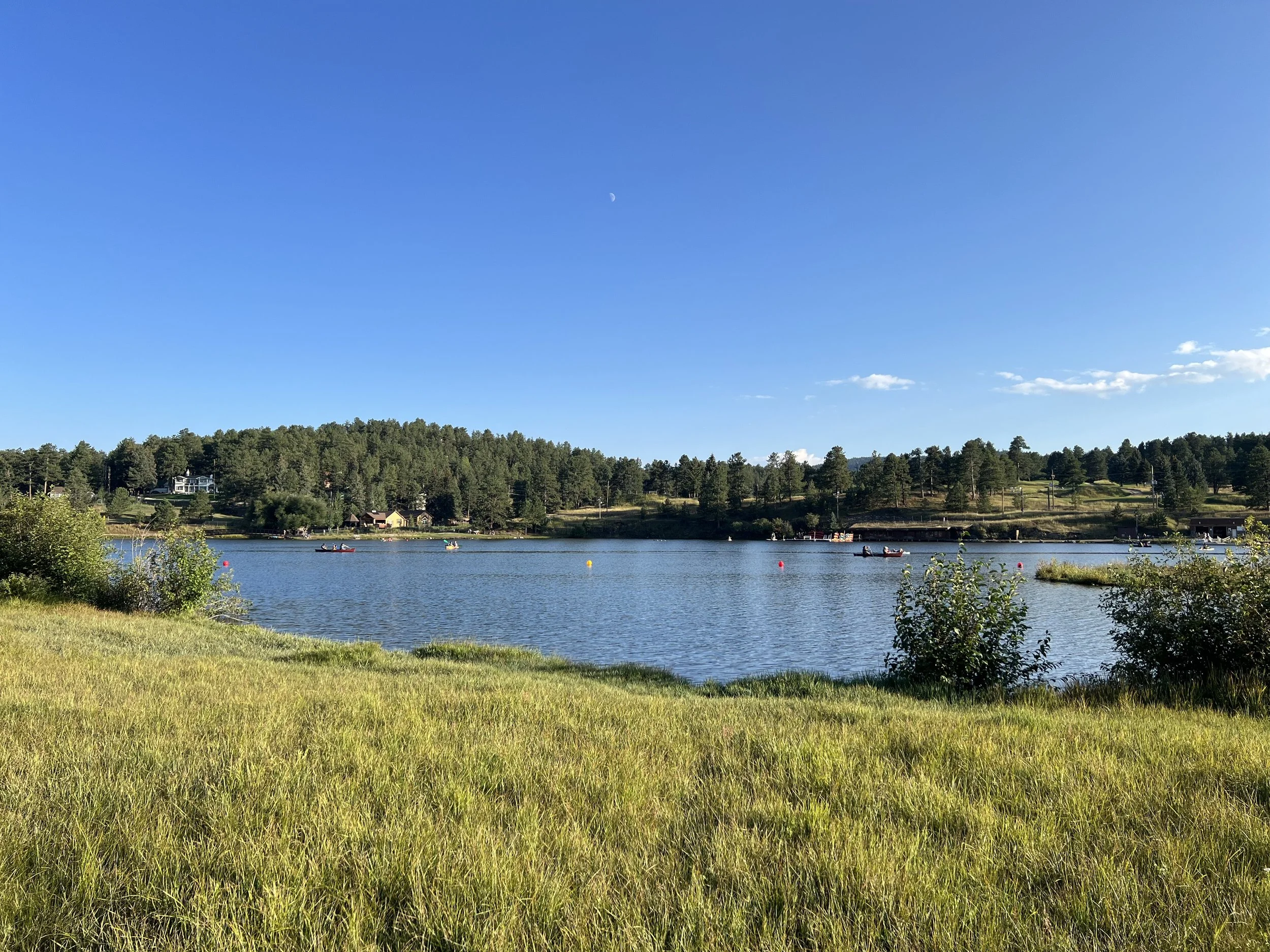Scenic view of a lake with boats, backed by a tree-lined hillside under a clear blue sky with a crescent moon.