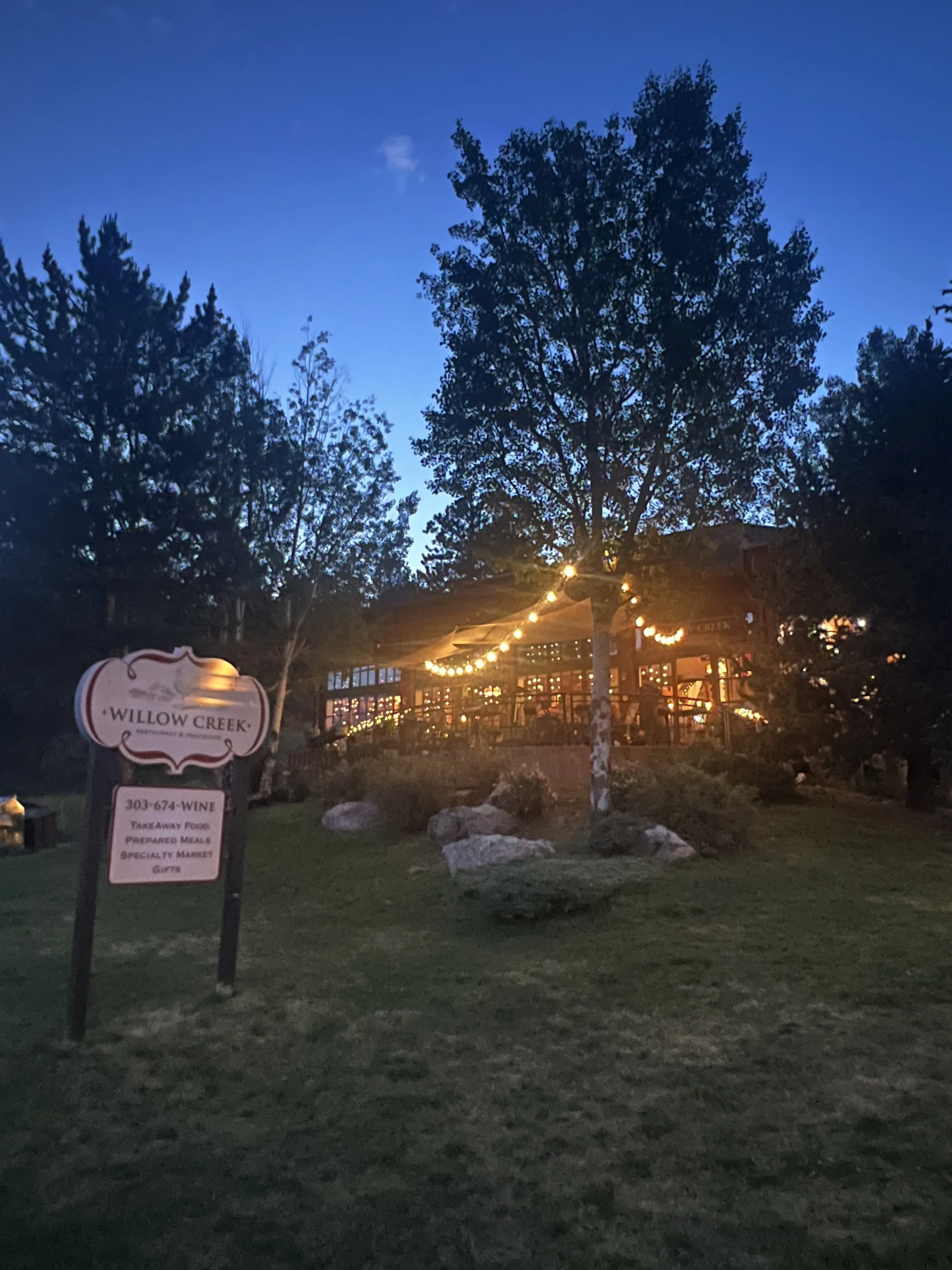 Outdoor dining area at Willow Creek restaurant illuminated with string lights at dusk, with trees surrounding the space and a sign in the foreground.