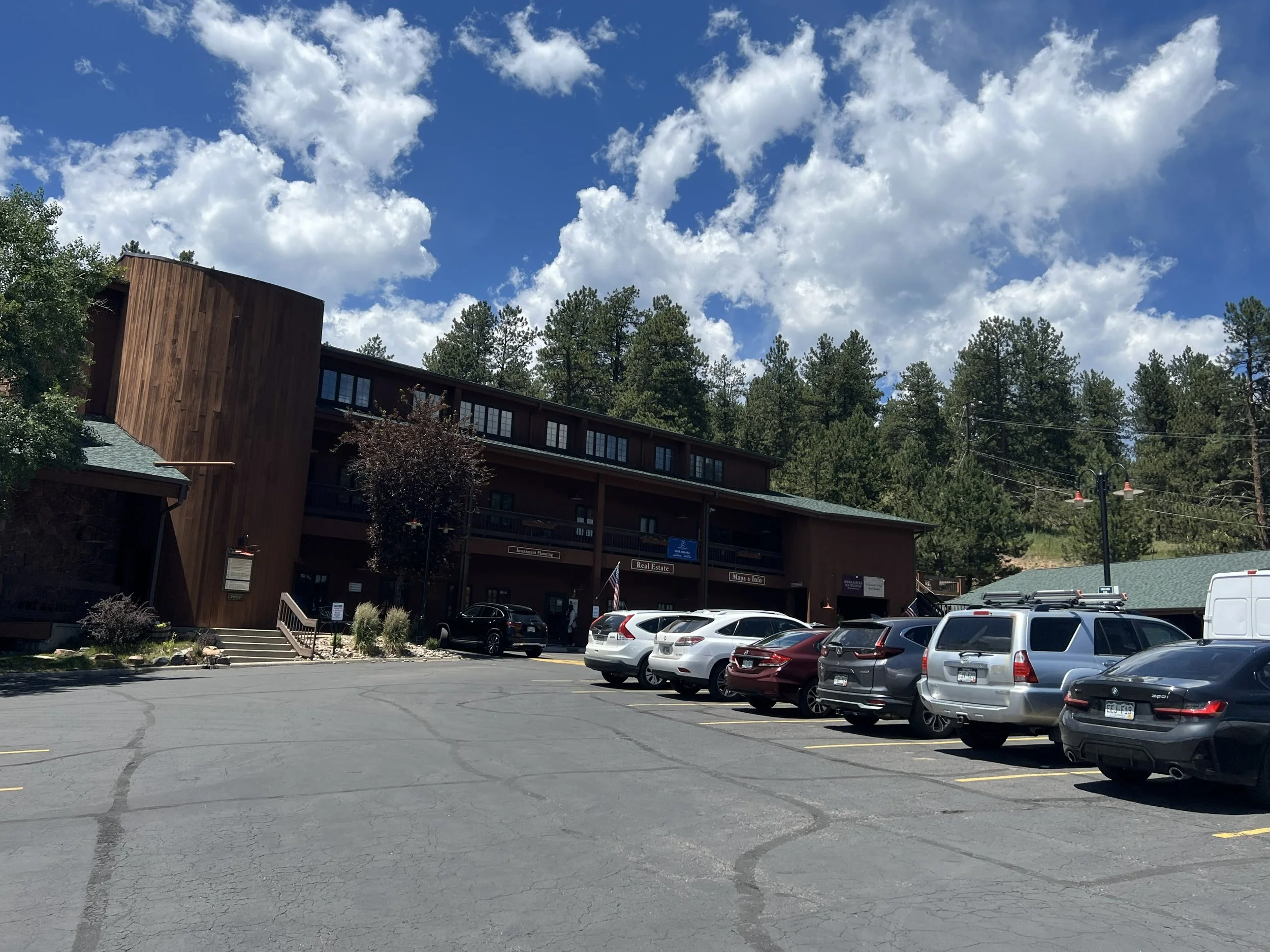 Multi-story wooden building with parked cars in front, surrounded by trees, under a partly cloudy sky.