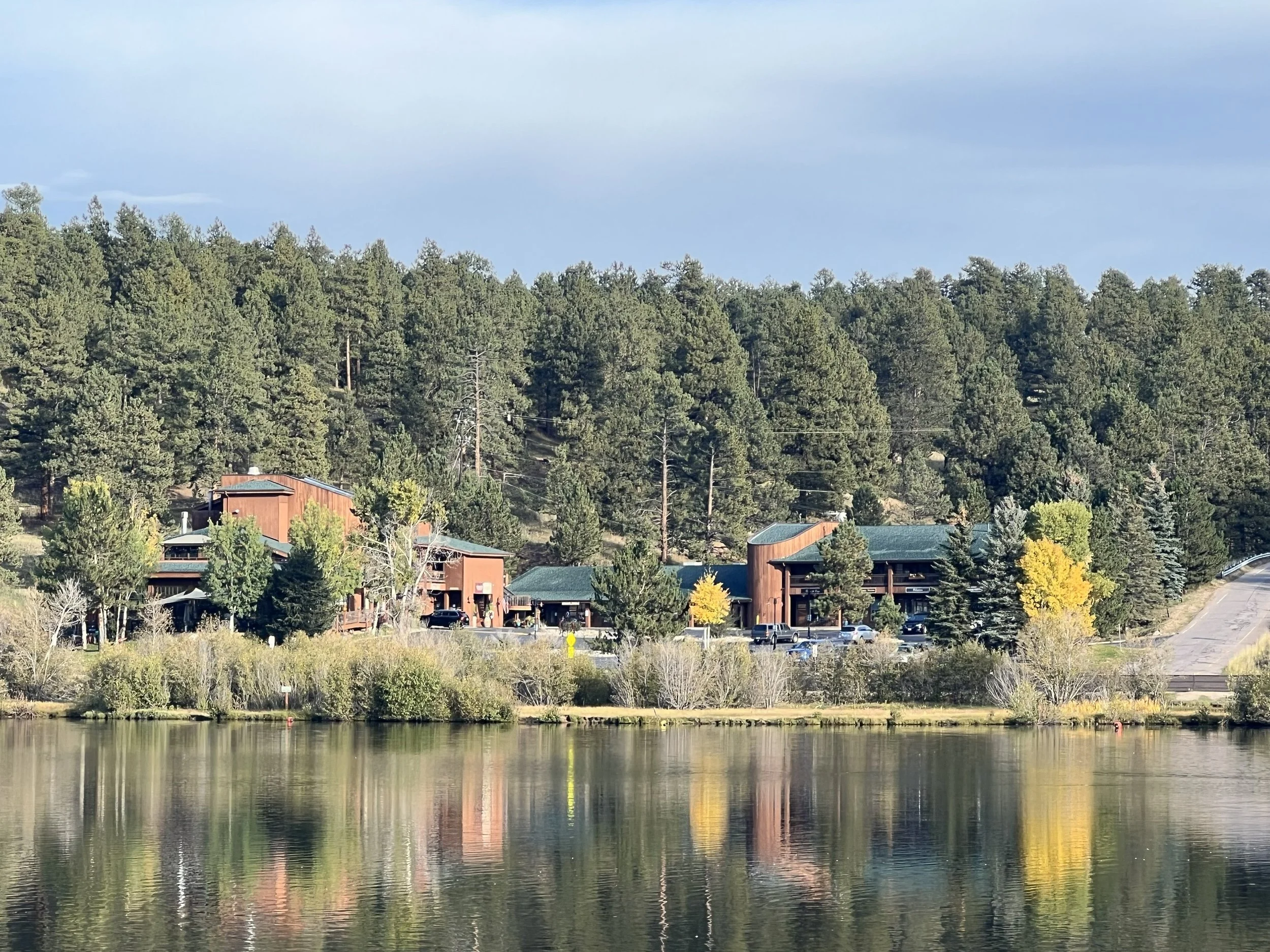A lakeside view of a wooded hillside with buildings and a parking lot, reflected in calm water.