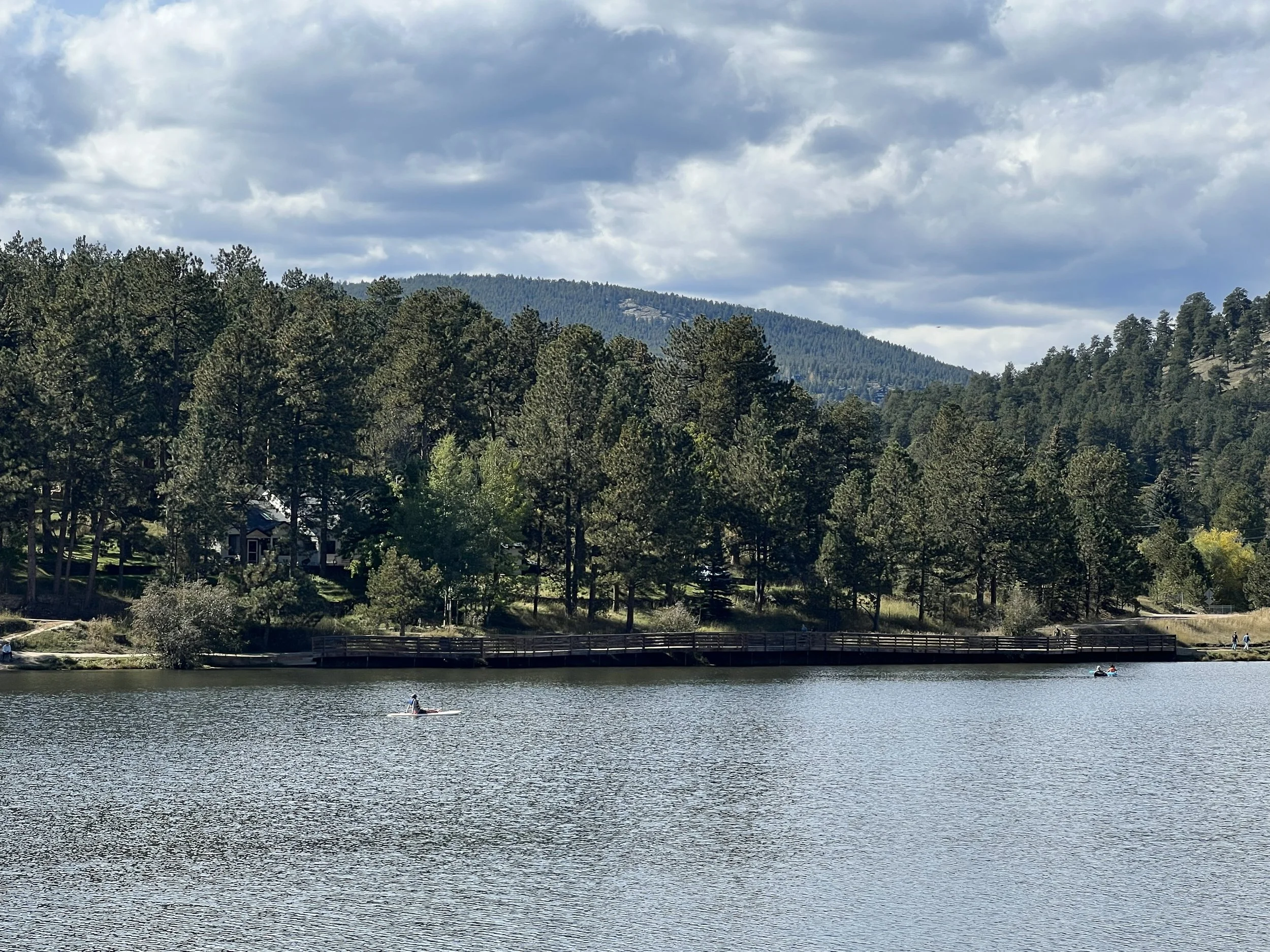 A lake with three people kayaking on the water, surrounded by a forested area with tall pine trees and hills in the background. There is a cloudy sky overhead.
