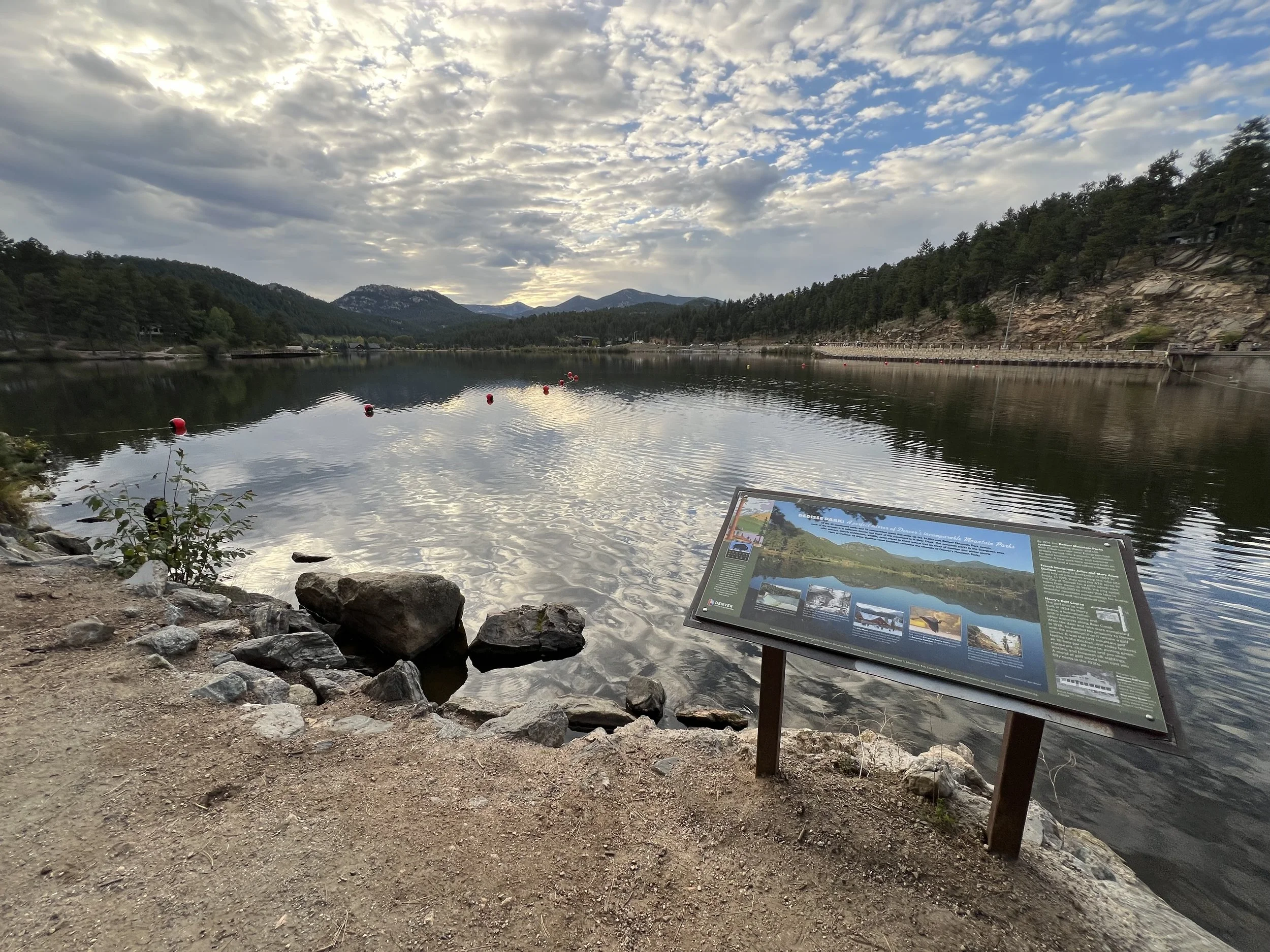 A peaceful lakeside scene with calm water reflecting the cloudy sky, distant mountains, and surrounding trees. There is an informational sign on the shore and orange buoys floating on the lake.