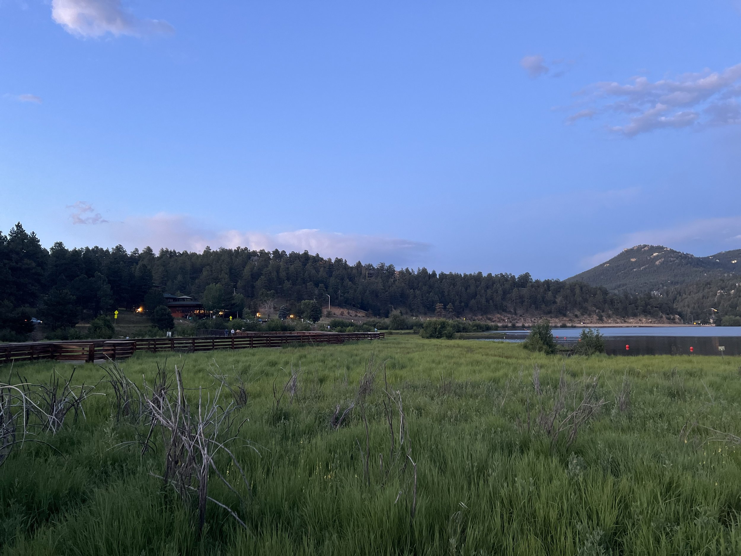 Scenic landscape with green grassy field in foreground, wooden fence, trees, lake, mountains in background, and partly cloudy sky with blue and gray clouds.