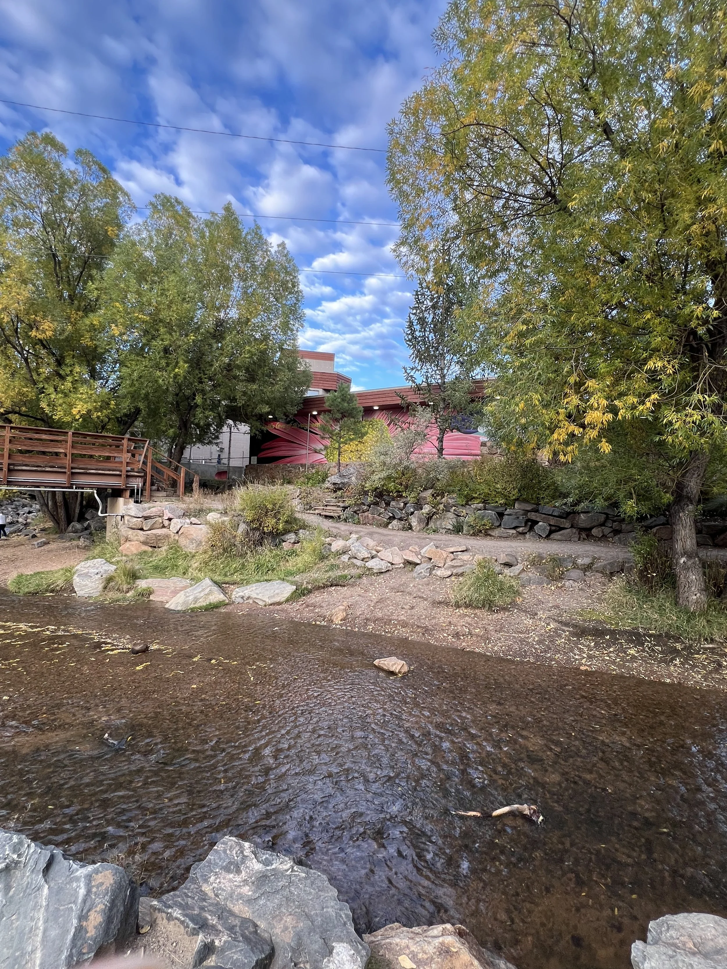 A small river or creek with rocks along the banks, surrounded by trees with fall foliage. There is a wooden walkway to the left and a building with a colorful mural in the background under a partly cloudy sky.