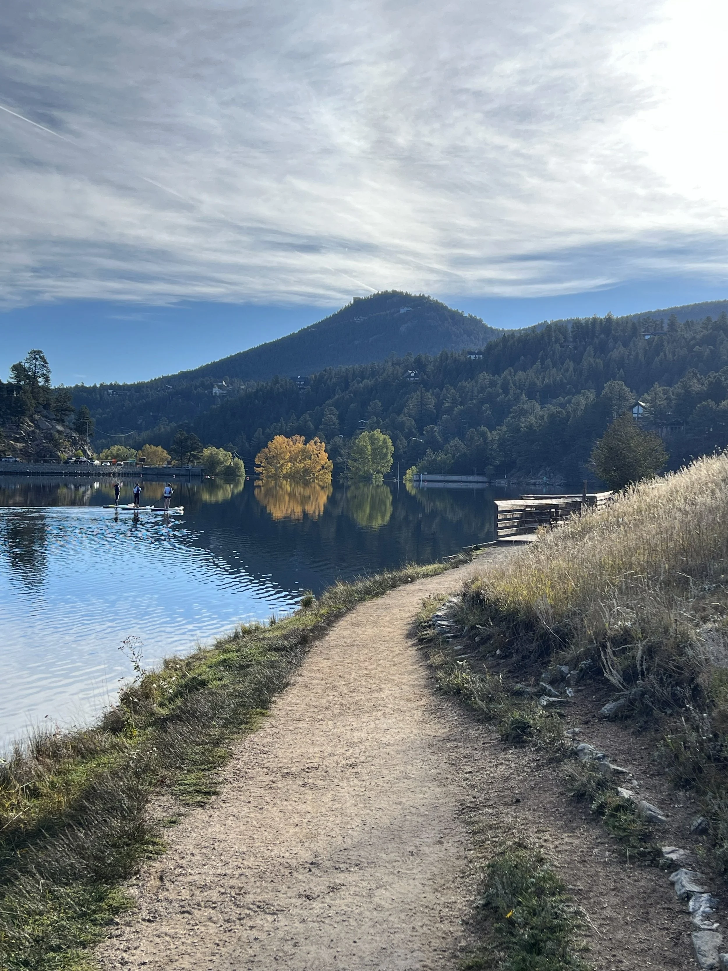 A dirt trail alongside a calm lake with trees reflecting in the water, surrounded by mountains and a partly cloudy sky.