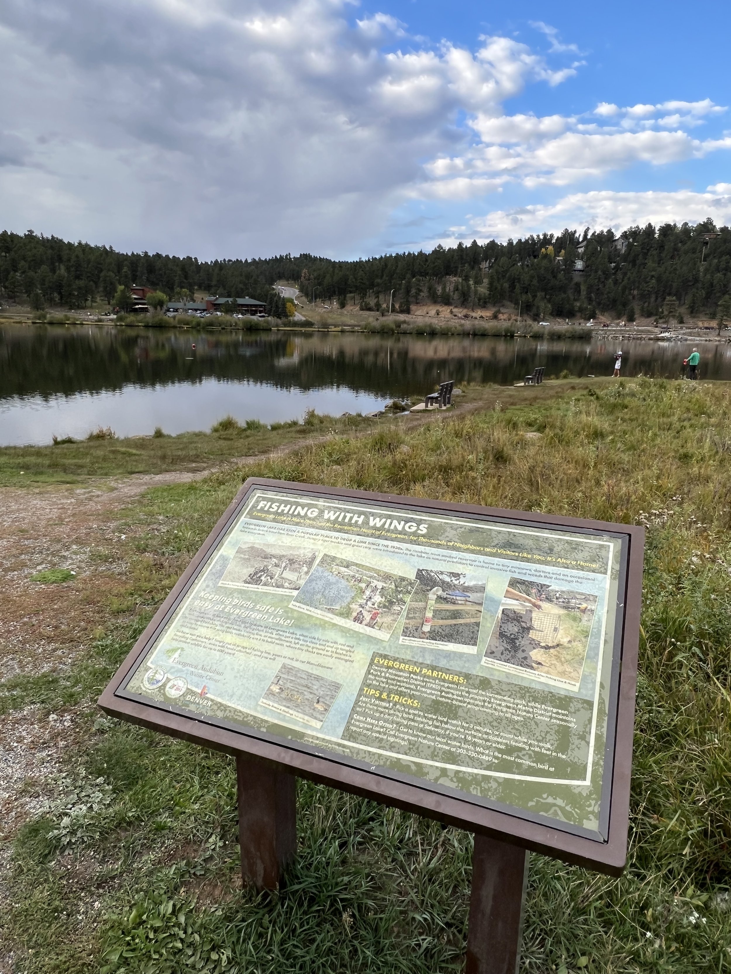 A lakeside scene with a signboard titled 'Fishing with Wings' in the foreground, three benches along the grassy shore, and people fishing in the calm lake. The background features a wooded hillside under a partly cloudy sky.
