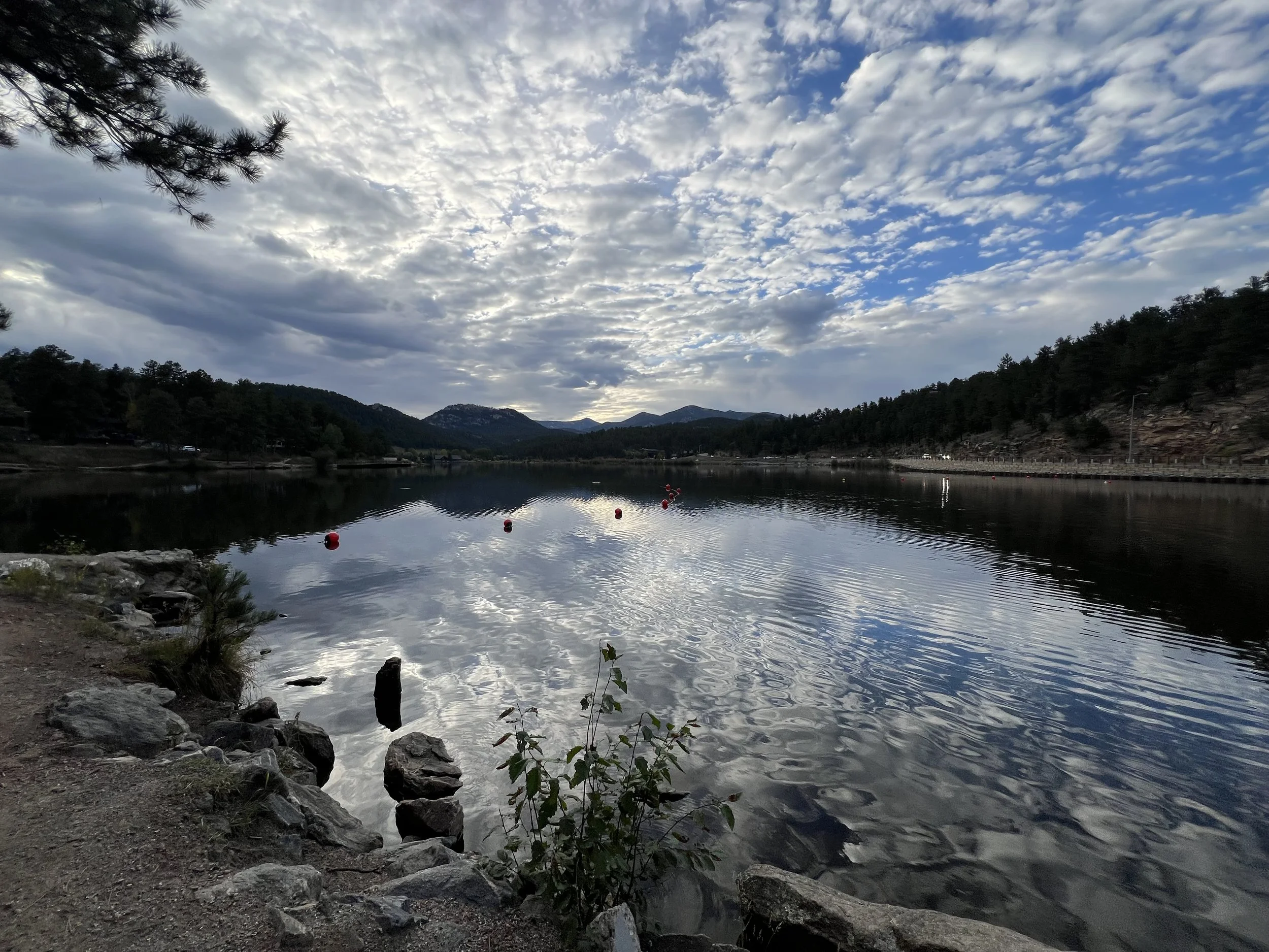 A peaceful lake surrounded by mountains and trees under a cloudy sky with some blue patches, reflecting the clouds and mountains on the water surface.