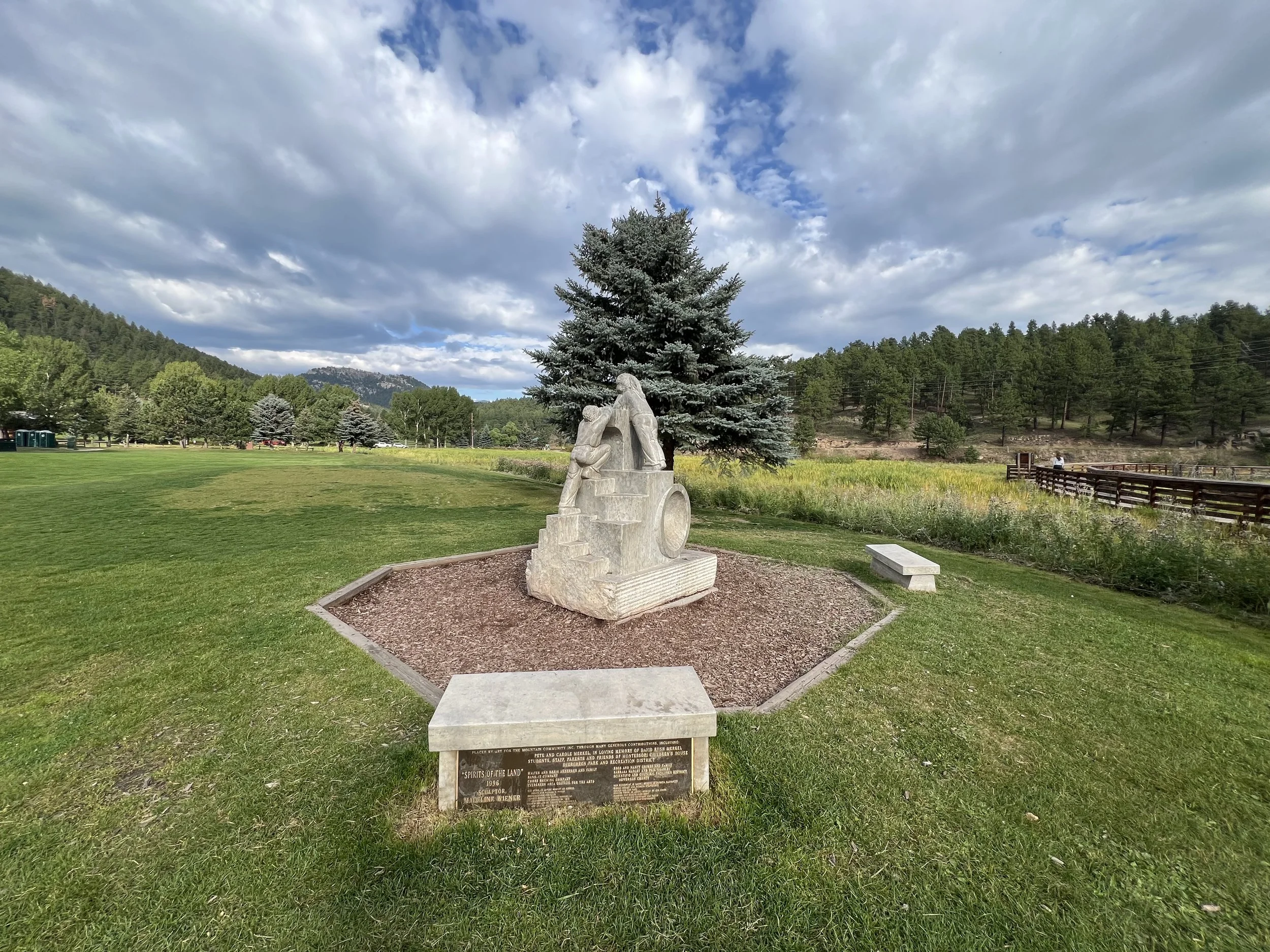 A sculpture of a person with children mounted on a stone base, located in a park with a grassy field, trees, mountains in the distance, and a partly cloudy sky.