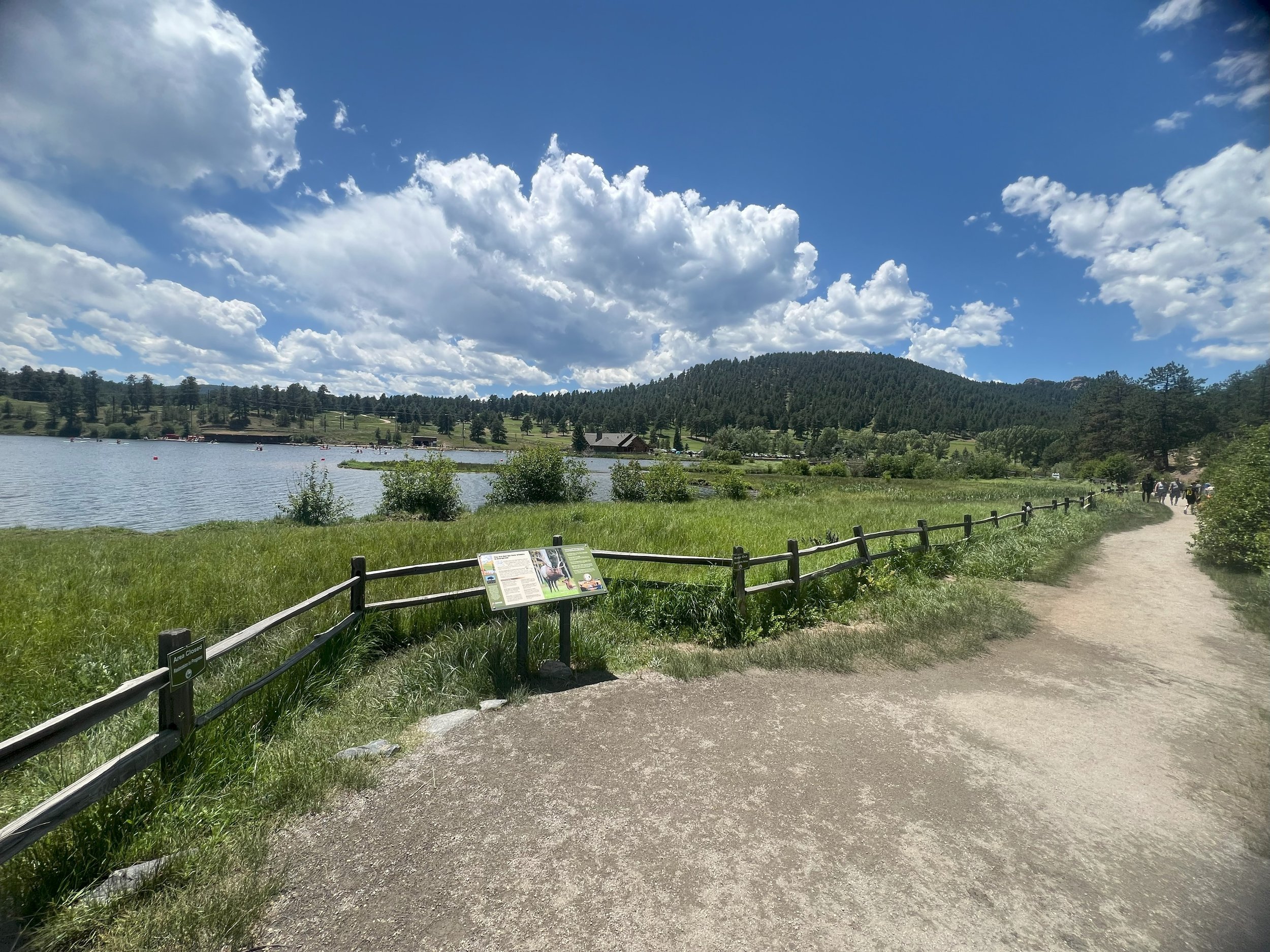 A scenic lake view with a gravel walking trail on the right, a wooden fence, green grass, trees, and a mountain under a partly cloudy blue sky.