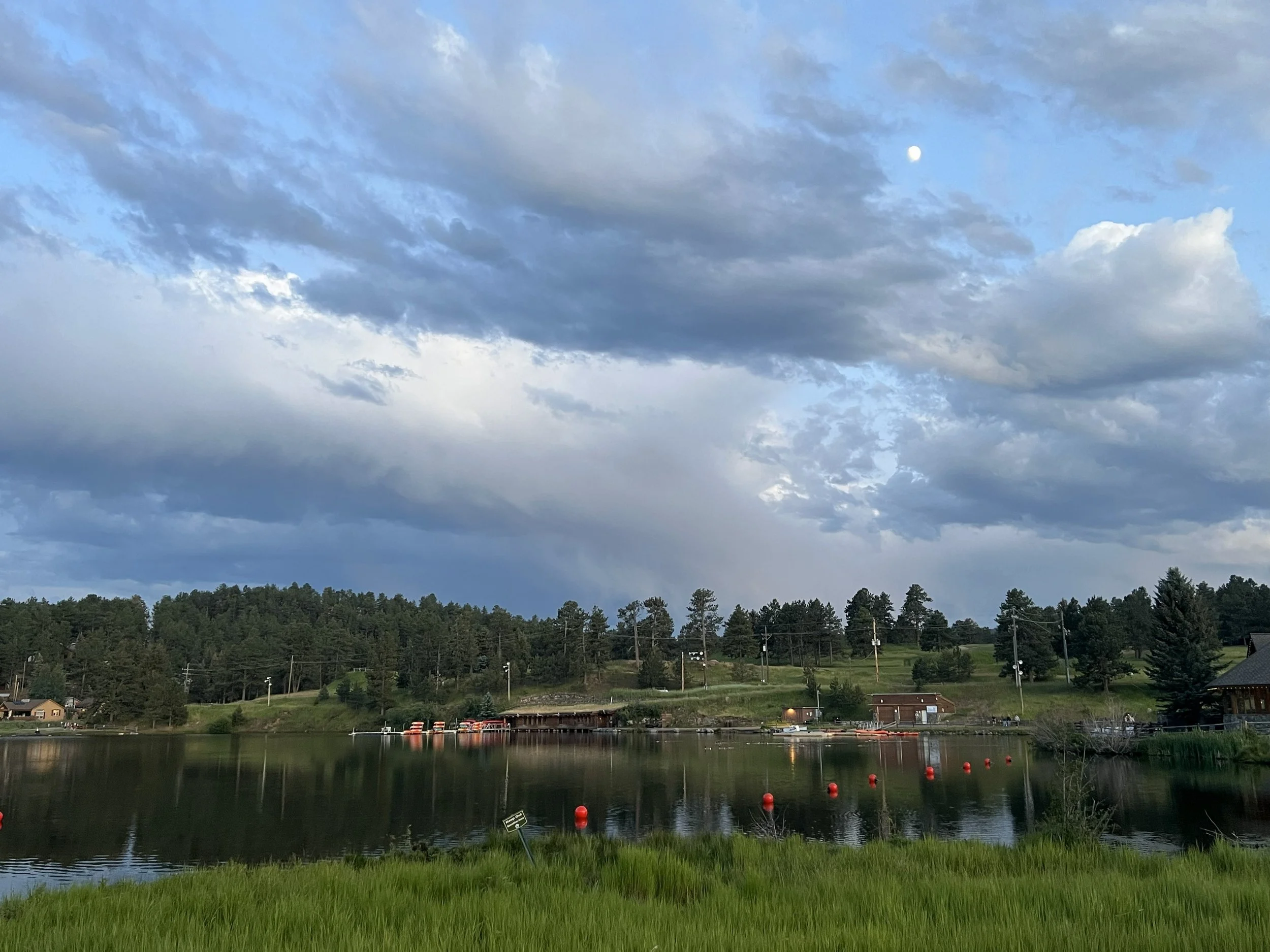 Scenic lake with green grass in foreground, calm water reflecting trees, small buildings, and red buoys, under a partly cloudy sky with the moon visible in the upper right.