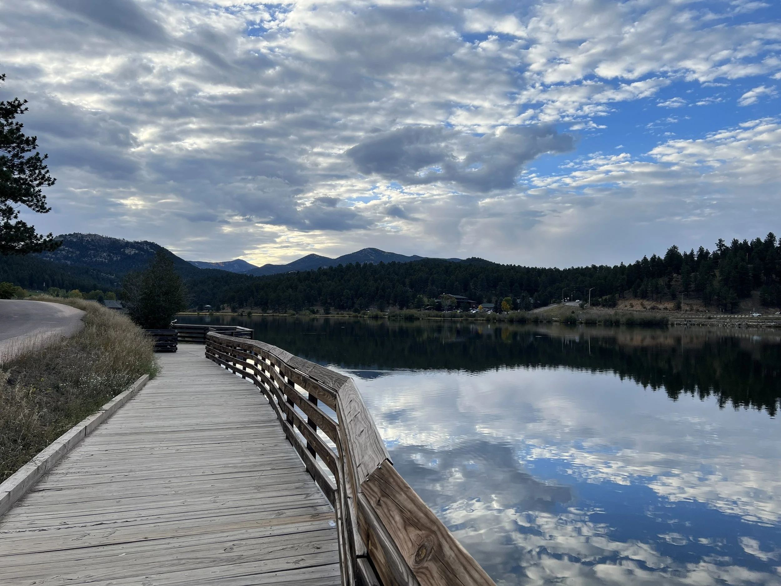 A wooden boardwalk along a lake with mountains in the distance and a cloudy sky reflected in the water.
