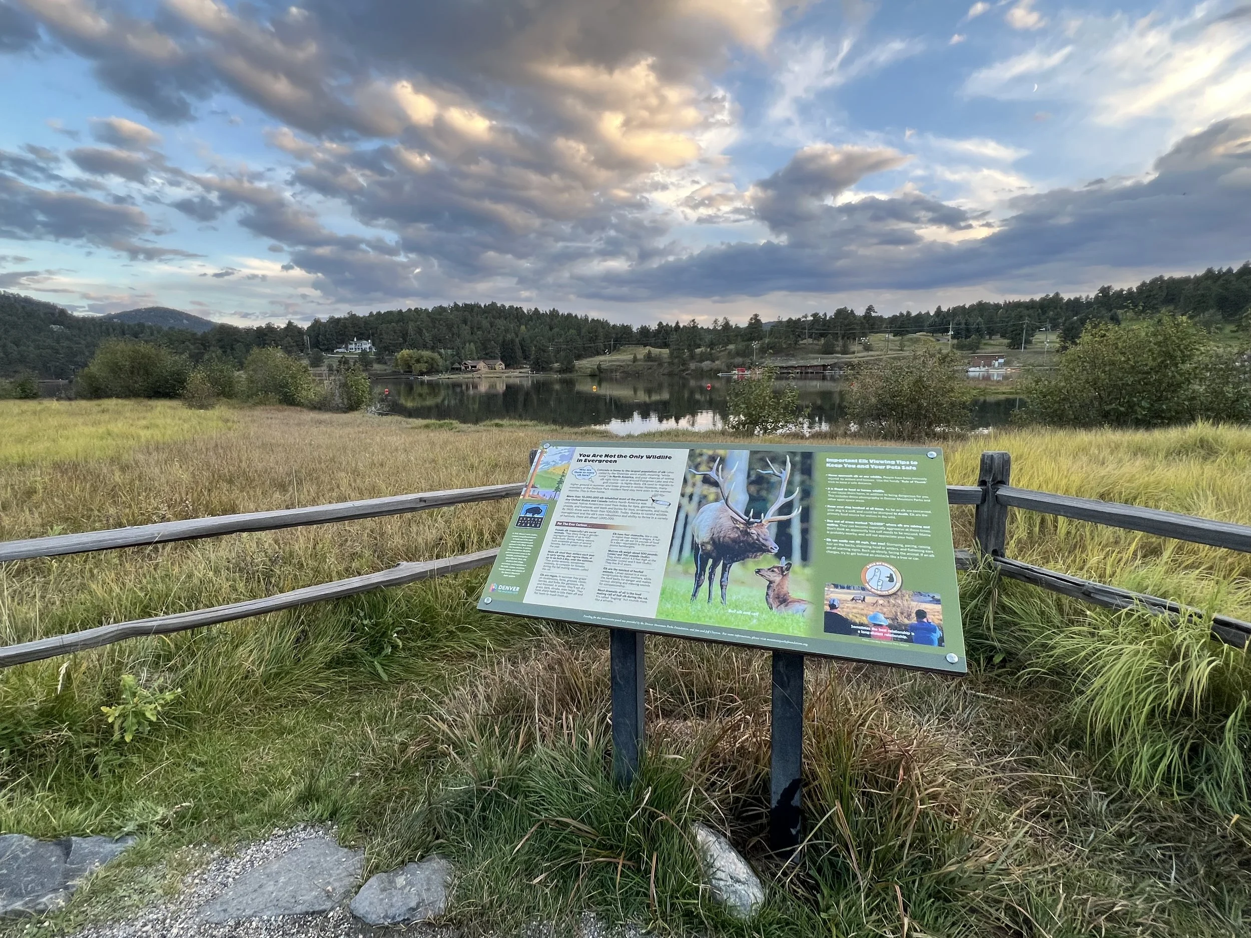 An informational sign near a scenic lake with grassy fields, trees, and mountains in the background under a partly cloudy sky.