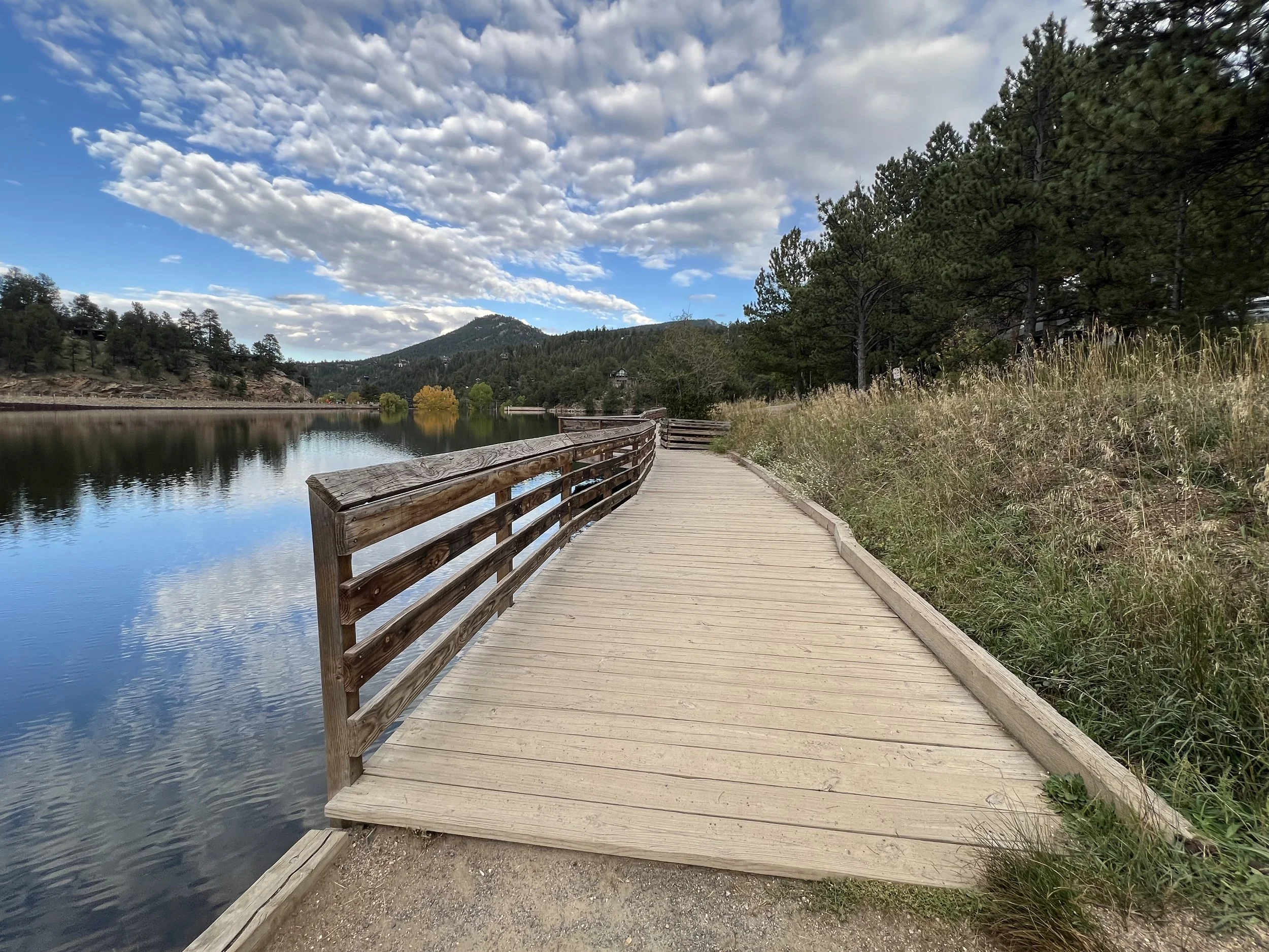 A wooden dock running alongside a calm lake with trees and mountains in the background under a partly cloudy sky.