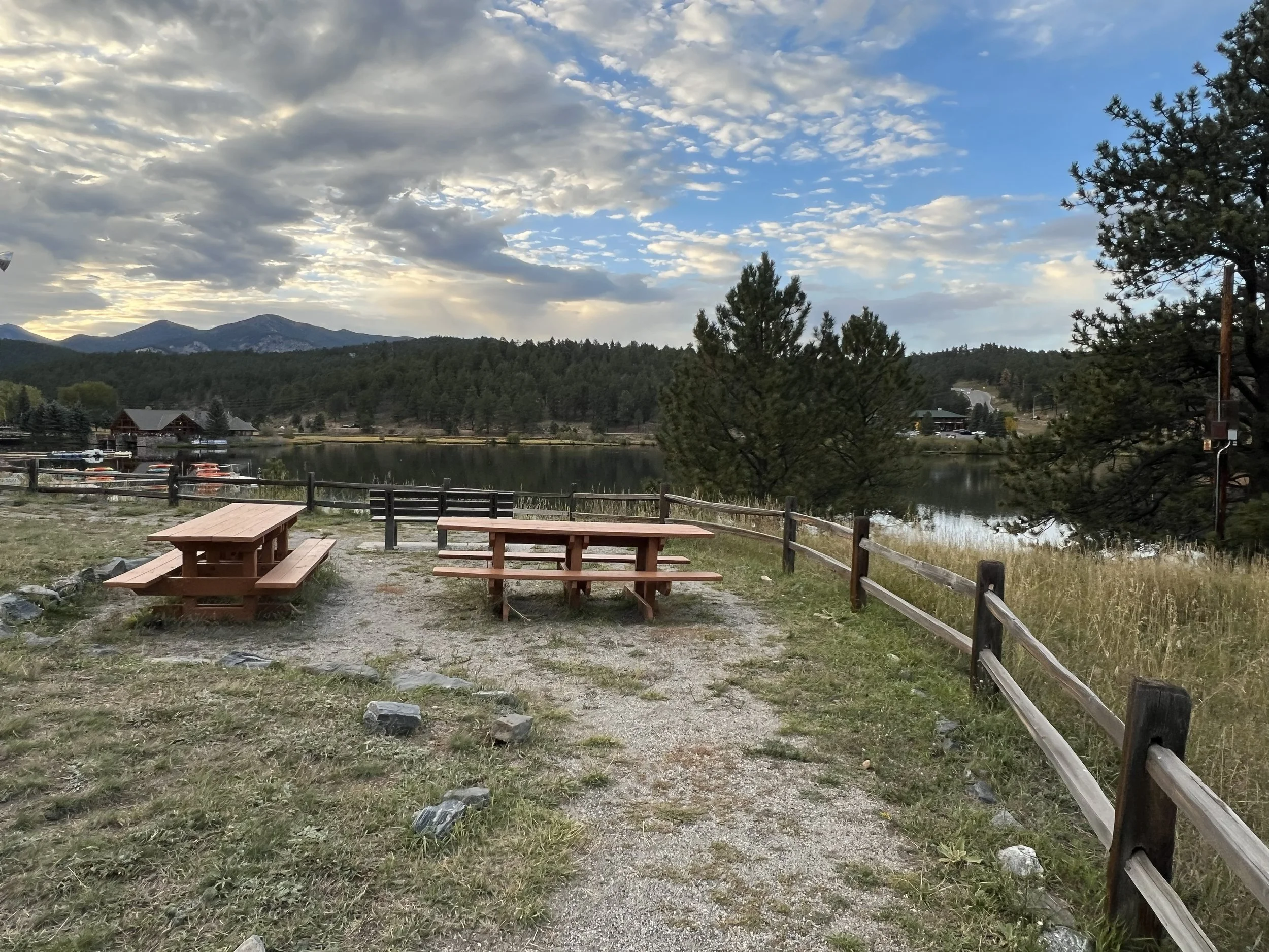 Two empty wooden picnic tables on a gravel and grass patch near a lake, with a wooden fence, trees, and mountains in the background under a partly cloudy sky.